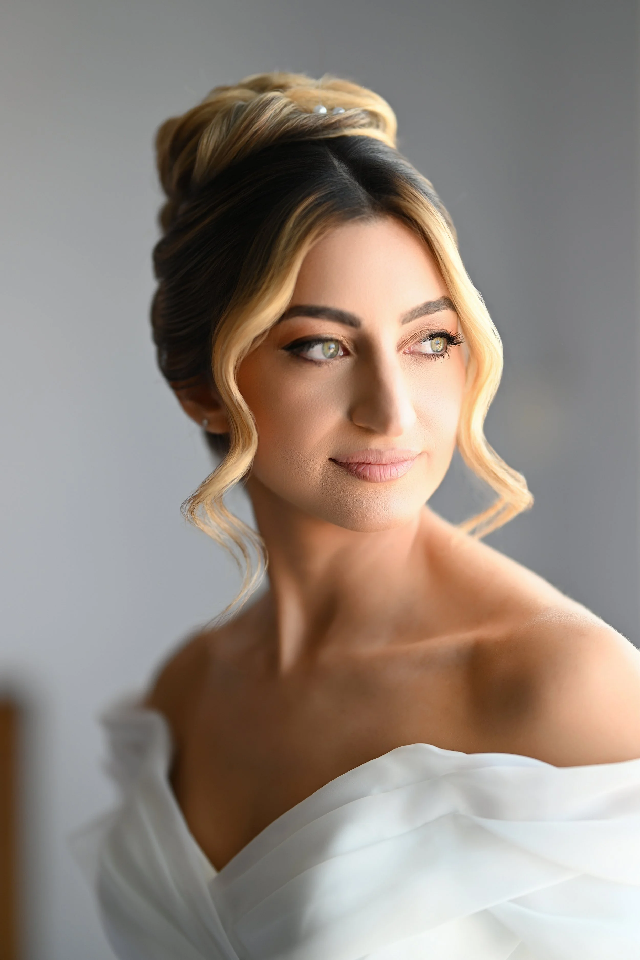 Close-up of a woman with styled blonde hair and green eyes, wearing a white off-shoulder dress.