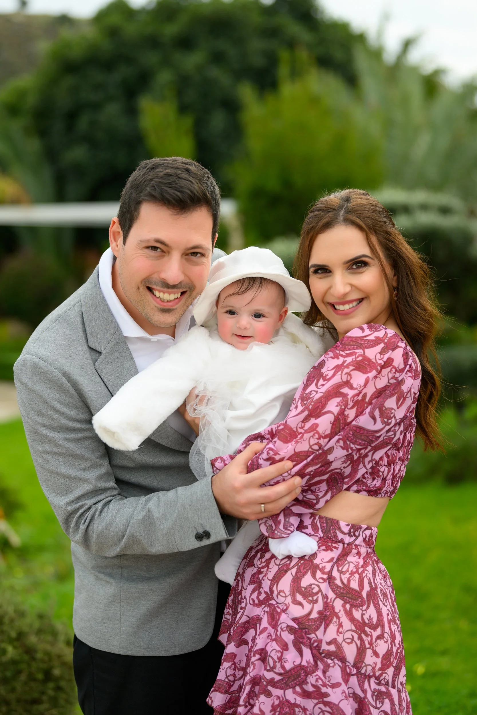 Smiling family of three outdoors: a man, woman, and baby in a garden with lush green trees.