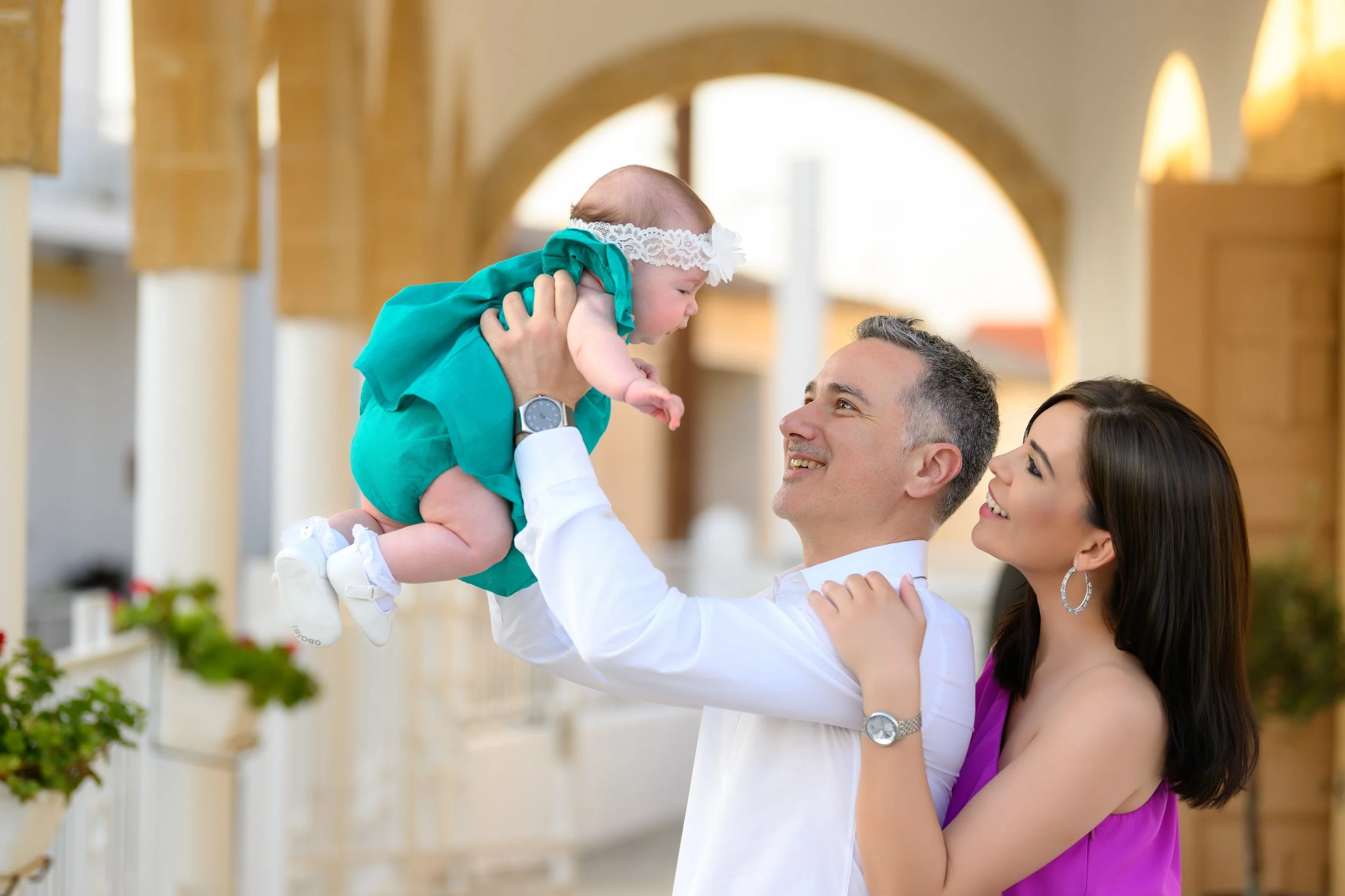 A man and woman smile at a baby girl the man is holding up. They are outdoors, in front of a building with arches, in sunny weather.