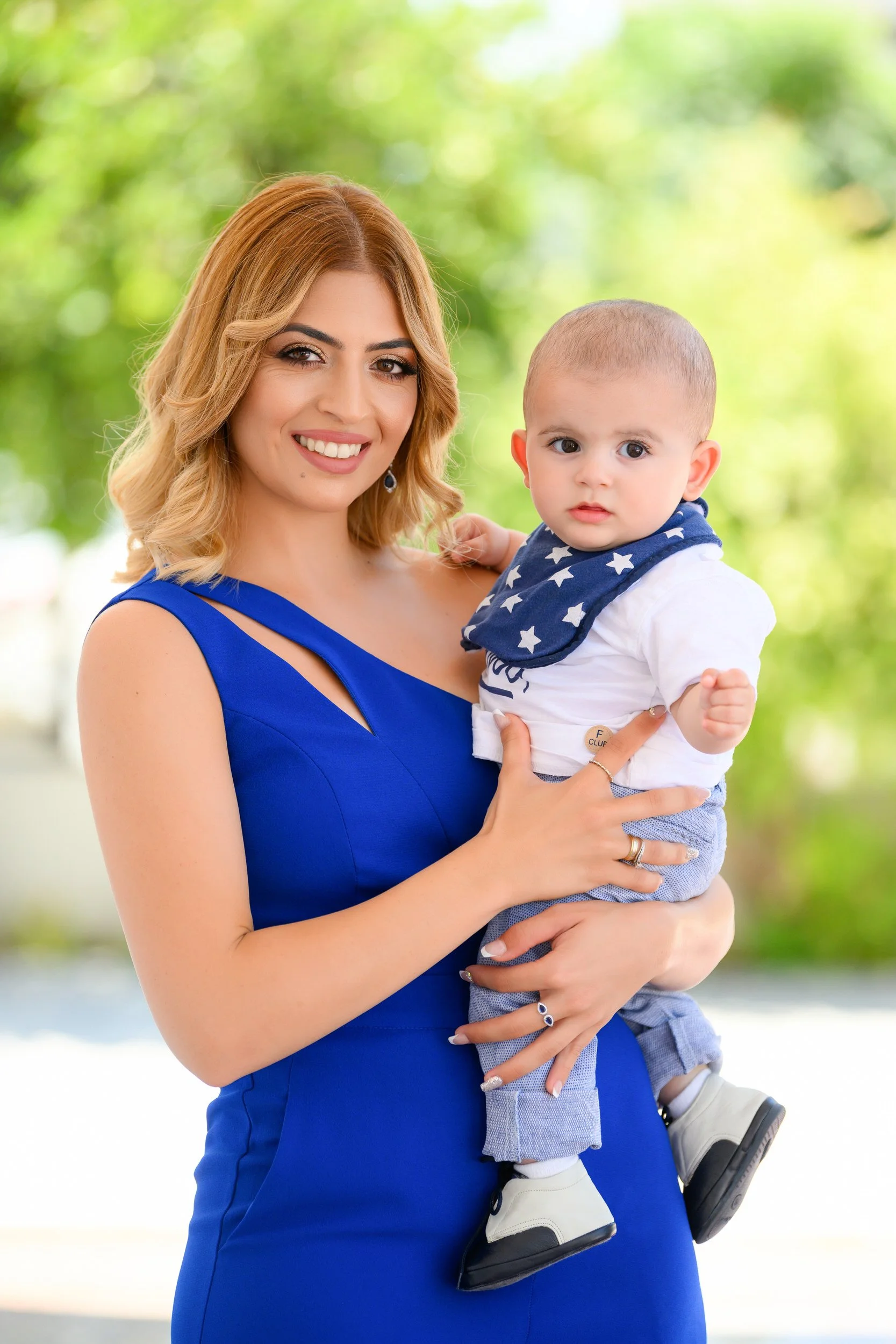 A woman with shoulder-length blonde hair in a blue dress holding a young boy with light skin, a crew cut, white shirt, blue patterned bandana, and light blue pants outside with green trees in the background.
