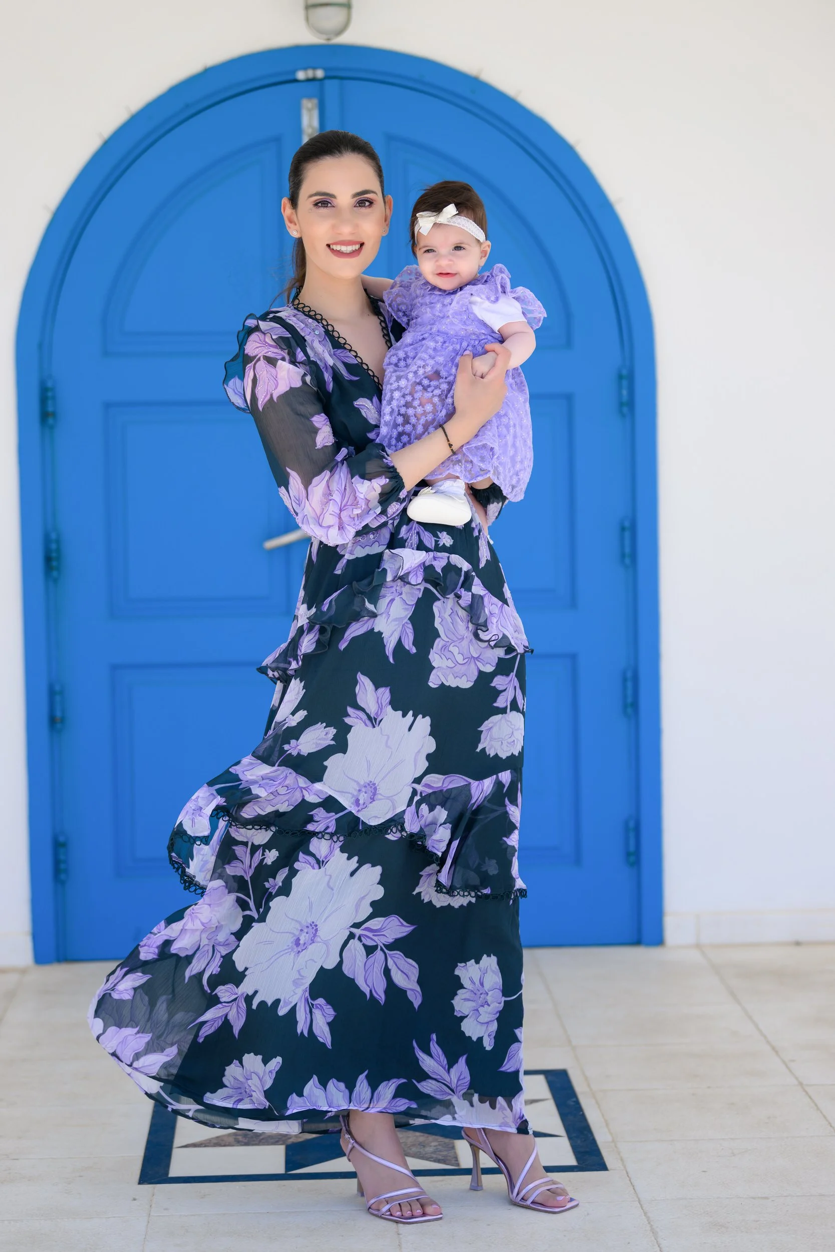 A woman in a black and purple floral dress holding a smiling toddler girl in a purple dress with a white headband, standing in front of a bright blue arched door.