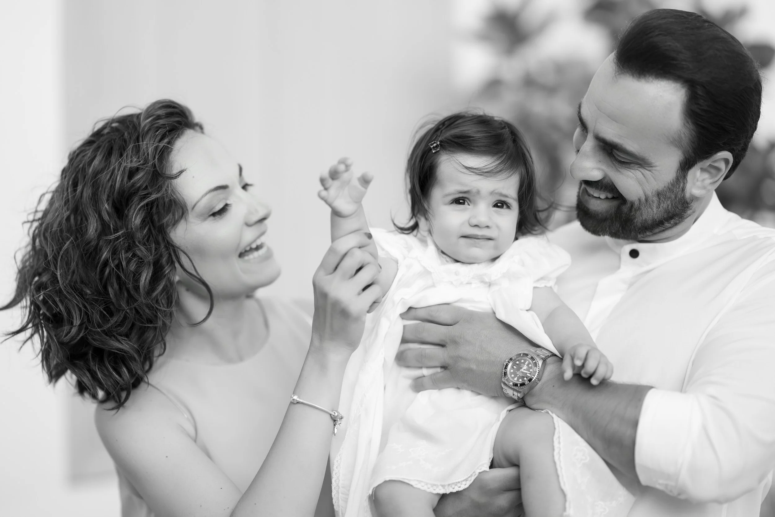 A family with a woman, a man, and a young girl, with the girl making a face while being held and supported by the man, during a joyful moment.