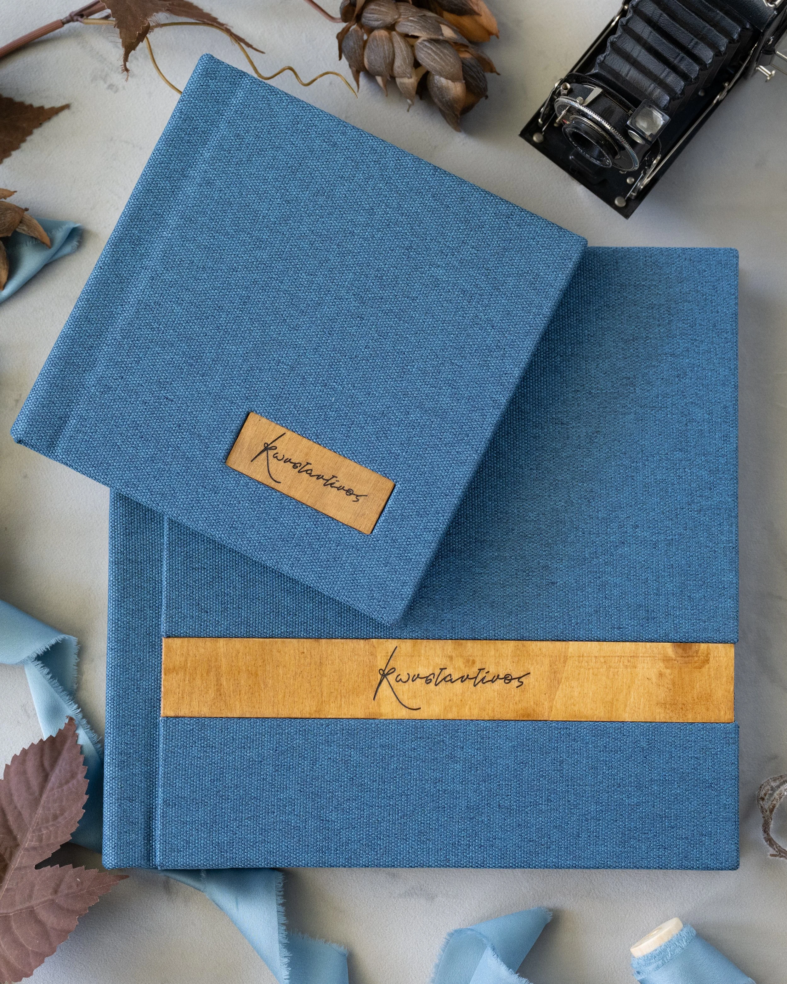 A blue fabric-covered lay flat album and matching folder with 'Konstantinos' written on labels, surrounded by dried leaves, a vintage camera, and fabric swatches.