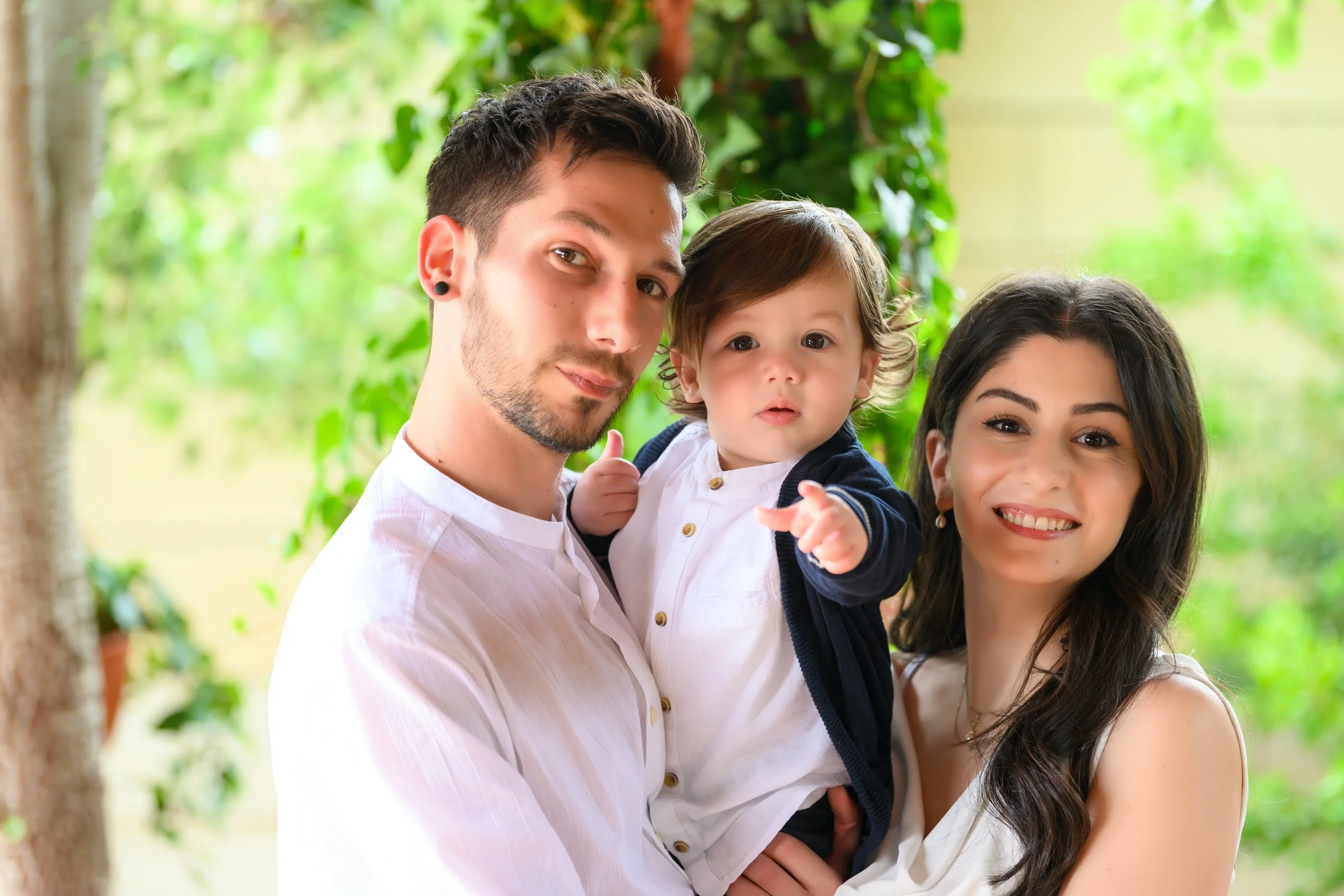 A happy family of three posing outdoors with green foliage in the background. A man with dark hair and a woman with long dark hair hold their young child, who is pointing at the camera.