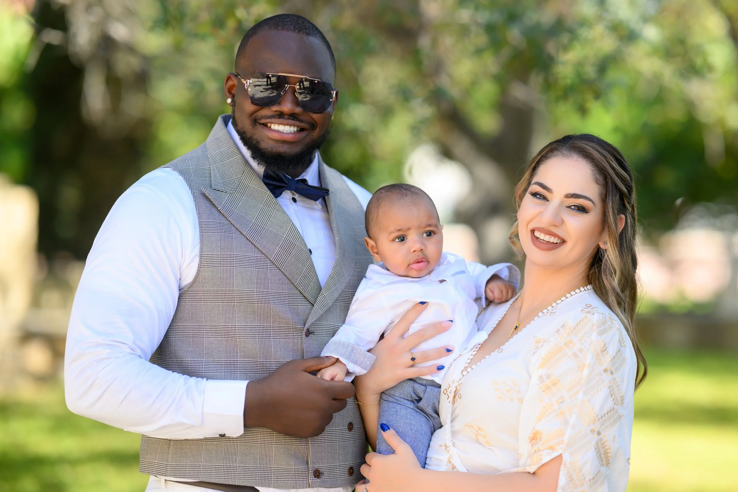 A family of three outdoors including a smiling man wearing sunglasses, a woman holding a baby, both smiling, with trees in the background.