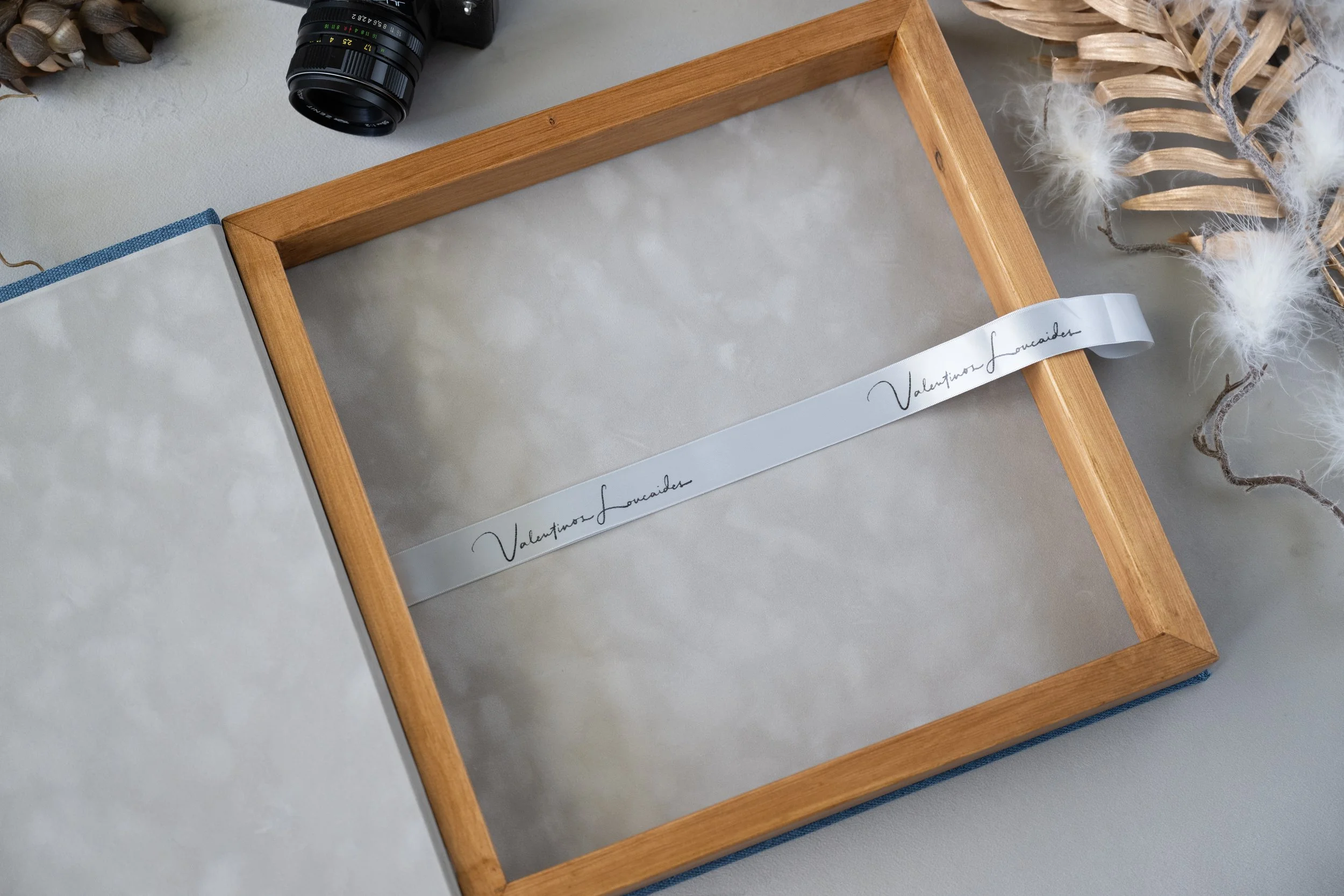 Empty luxury wooden album box with a white ribbon that has 'Valentino Loucaides' written on it, surrounded by a camera lens, dried flowers, and decorative white fuzzy branches.