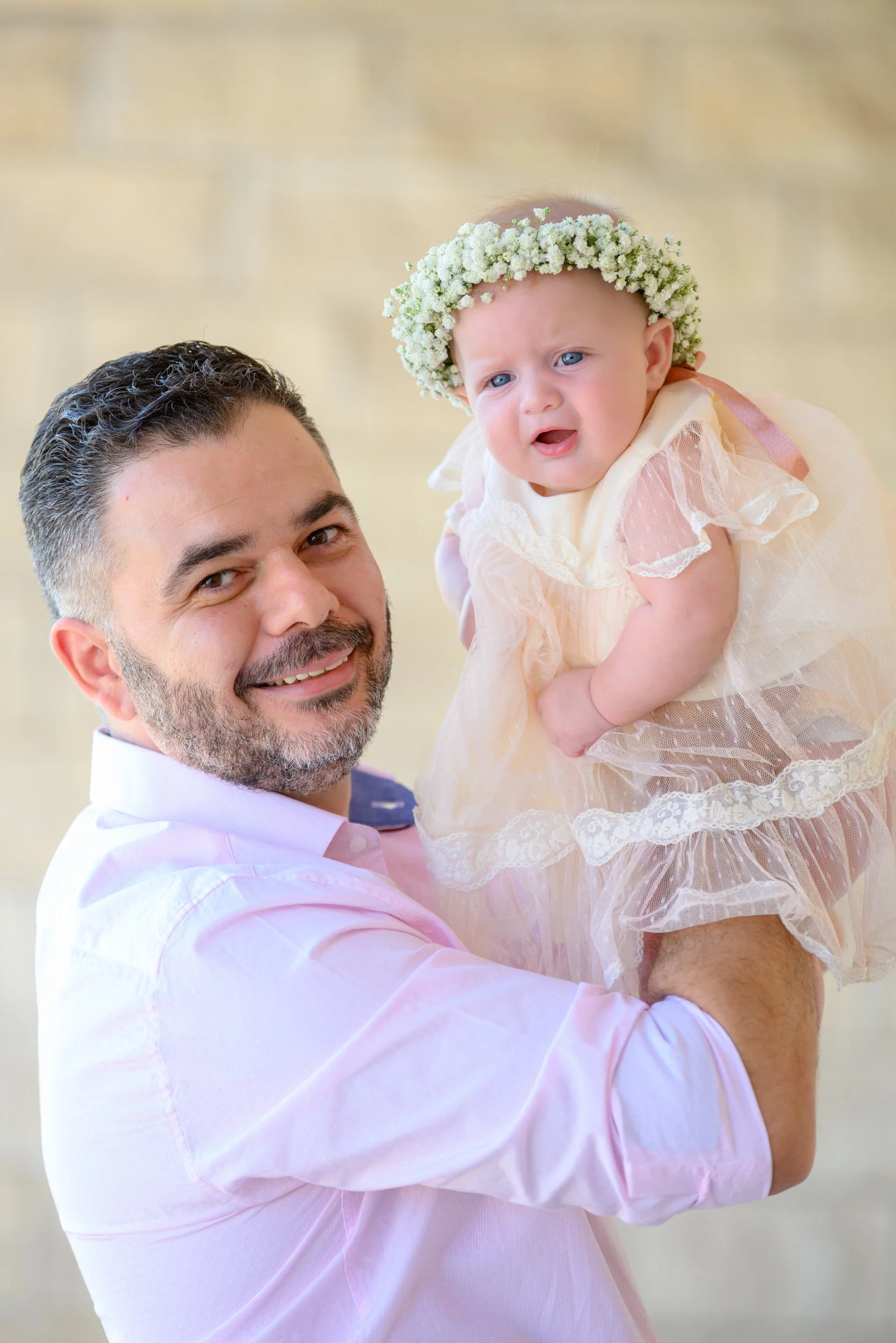 A man holding a baby girl wearing a floral headband and a lacy dress, smiling outdoors.