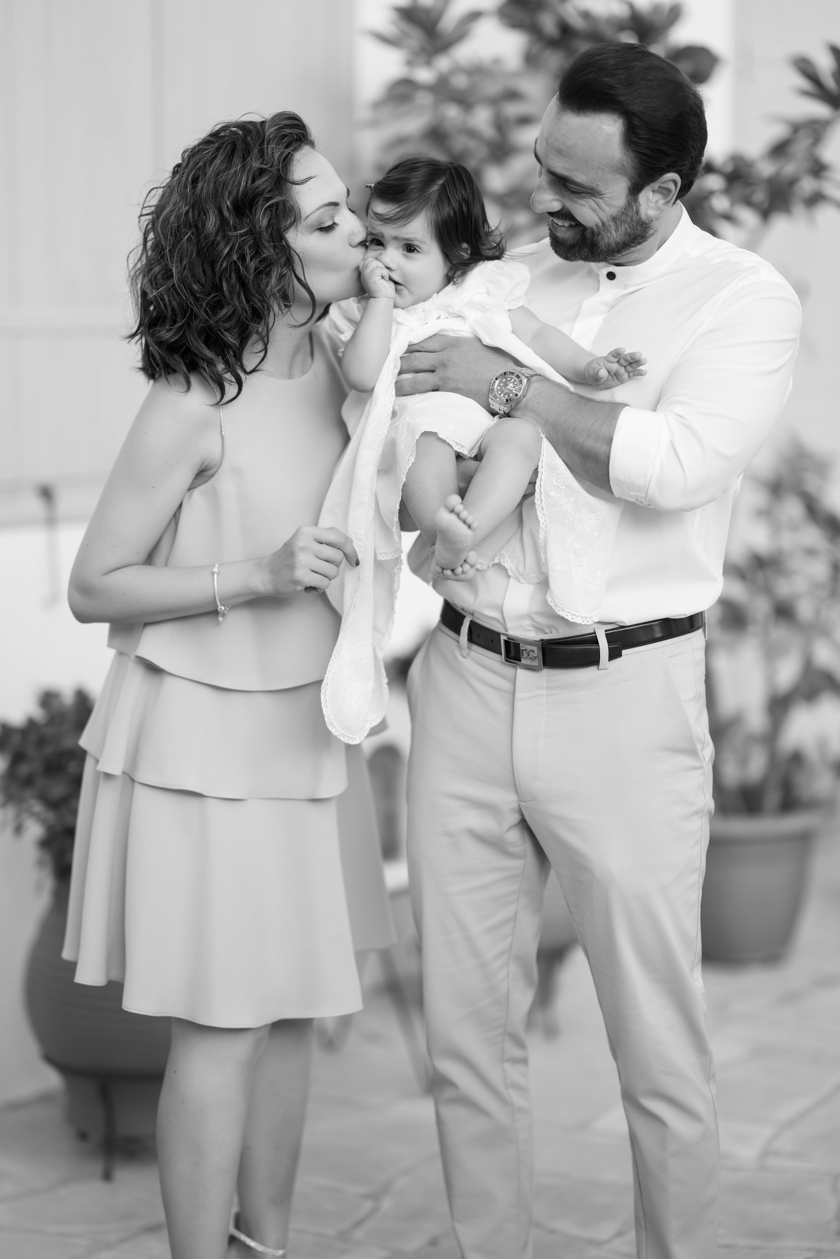 A family of three, a woman, a man, and a young girl, sharing a moment indoors. The woman is kissing the girl on the cheek, and the man is holding the girl, smiling at her. The photo is in black and white.