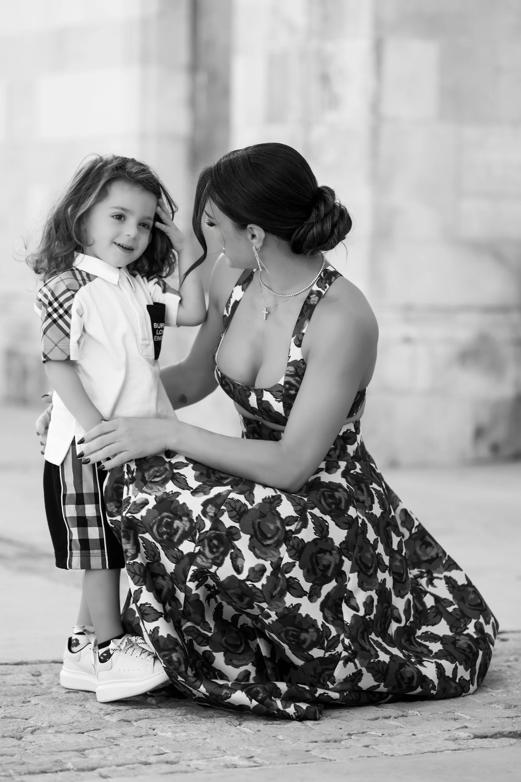 A woman with dark hair in an updo, wearing a floral dress, kneeling and smiling at a young girl with curly hair, who is touching her forehead and also smiling. The girl is dressed in a sports outfit with plaid shorts and sneakers. They are on a cobbl