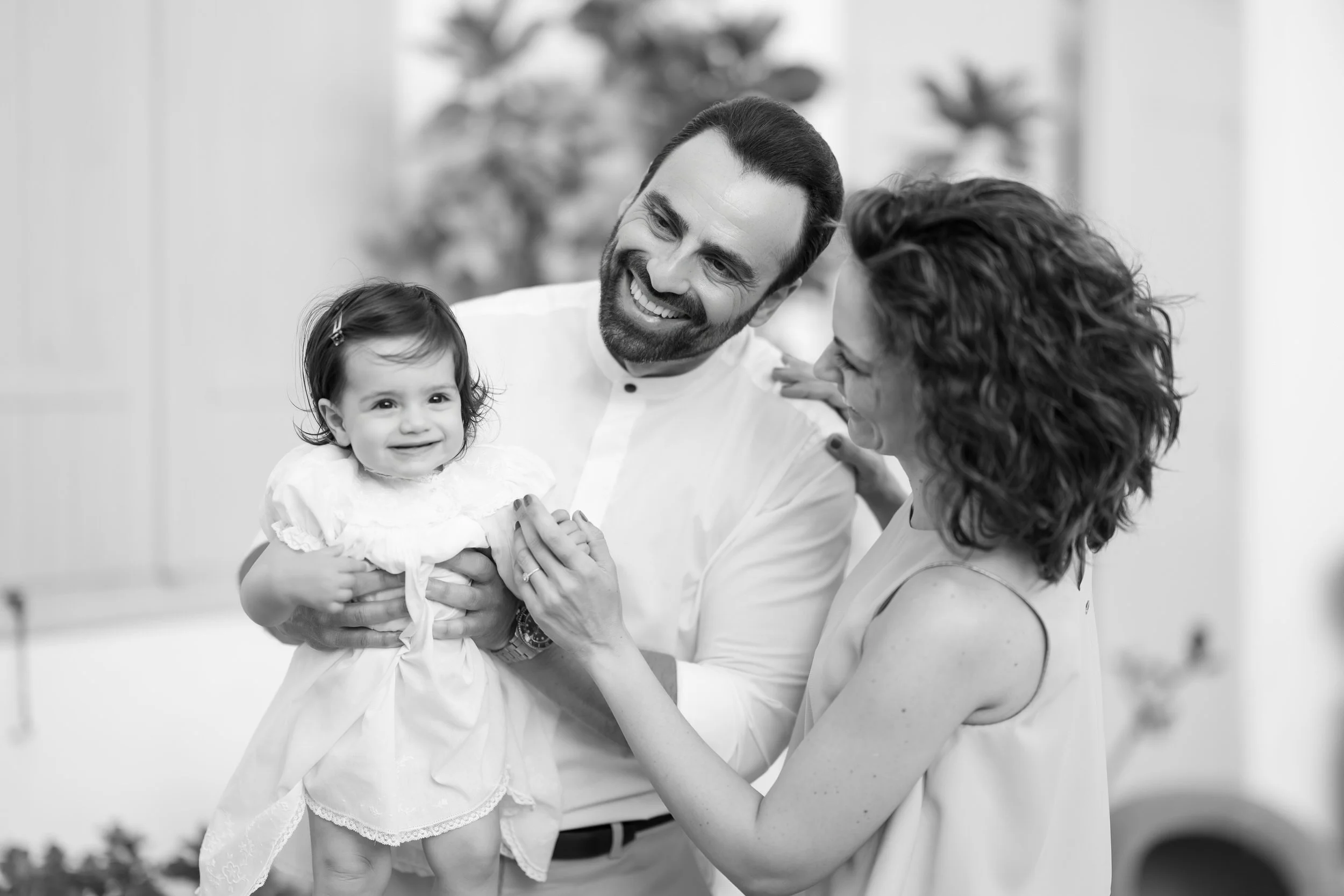 A smiling baby girl in a white dress held by a man and looking at a woman, all smiling and enjoying a happy moment together.