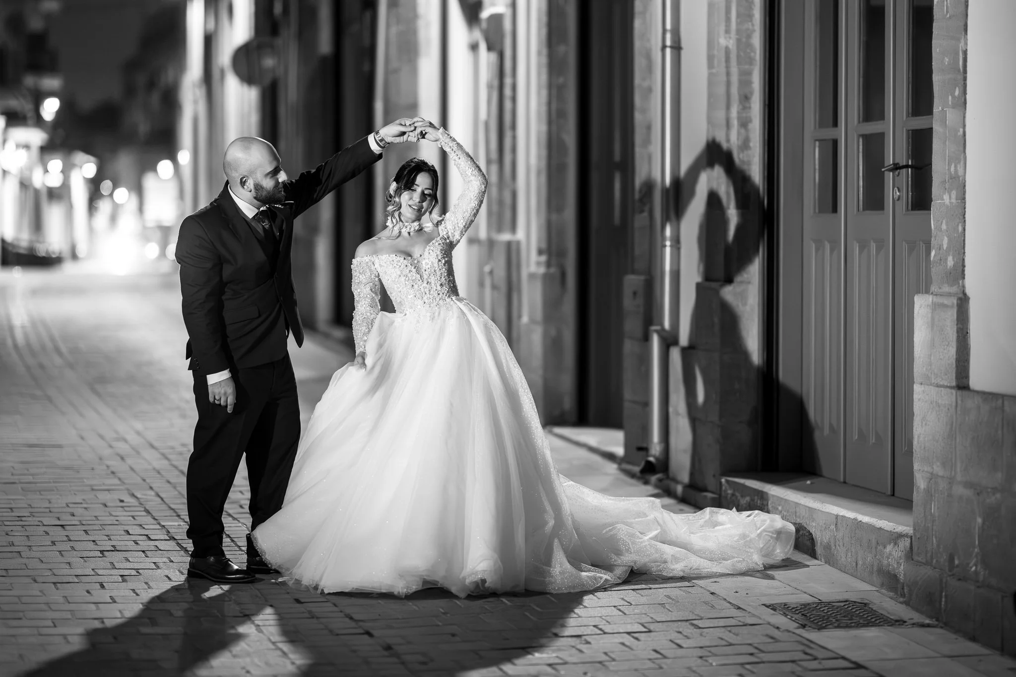 A couple dances on a city street at night, in wedding attire. The bride wears a lacy, off-shoulder wedding gown, and the groom wears a suit. They are holding hands in a twirl, with their shadows cast on a nearby wall.