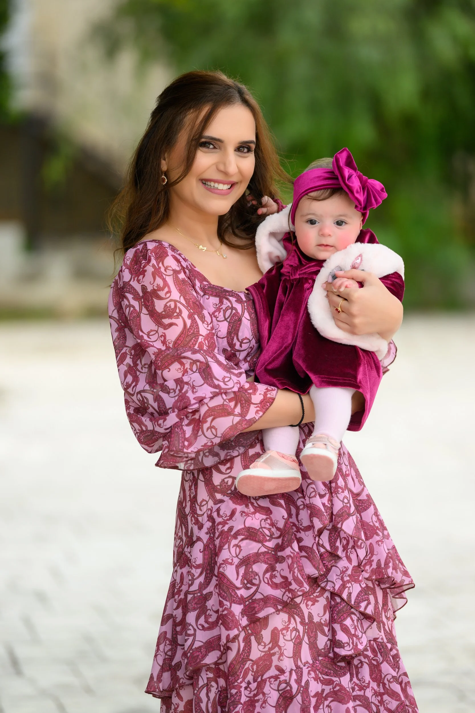 A smiling woman holding a baby girl outdoors with green trees in the background. The woman wears a pink and burgundy patterned dress, and the baby is dressed in a burgundy velvet outfit with a matching headband.