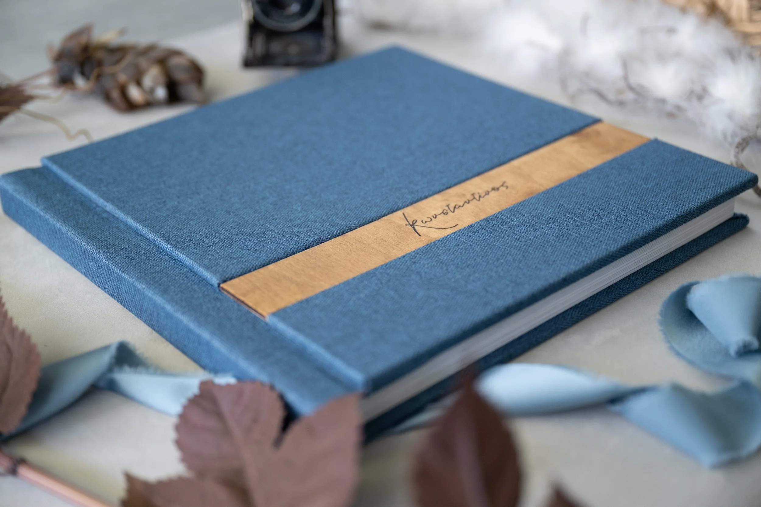 A blue hardcover lay flat album with a gold stripe and the word 'fourestiotes' handwritten on it, surrounded by dried leaves and fabric flowers.