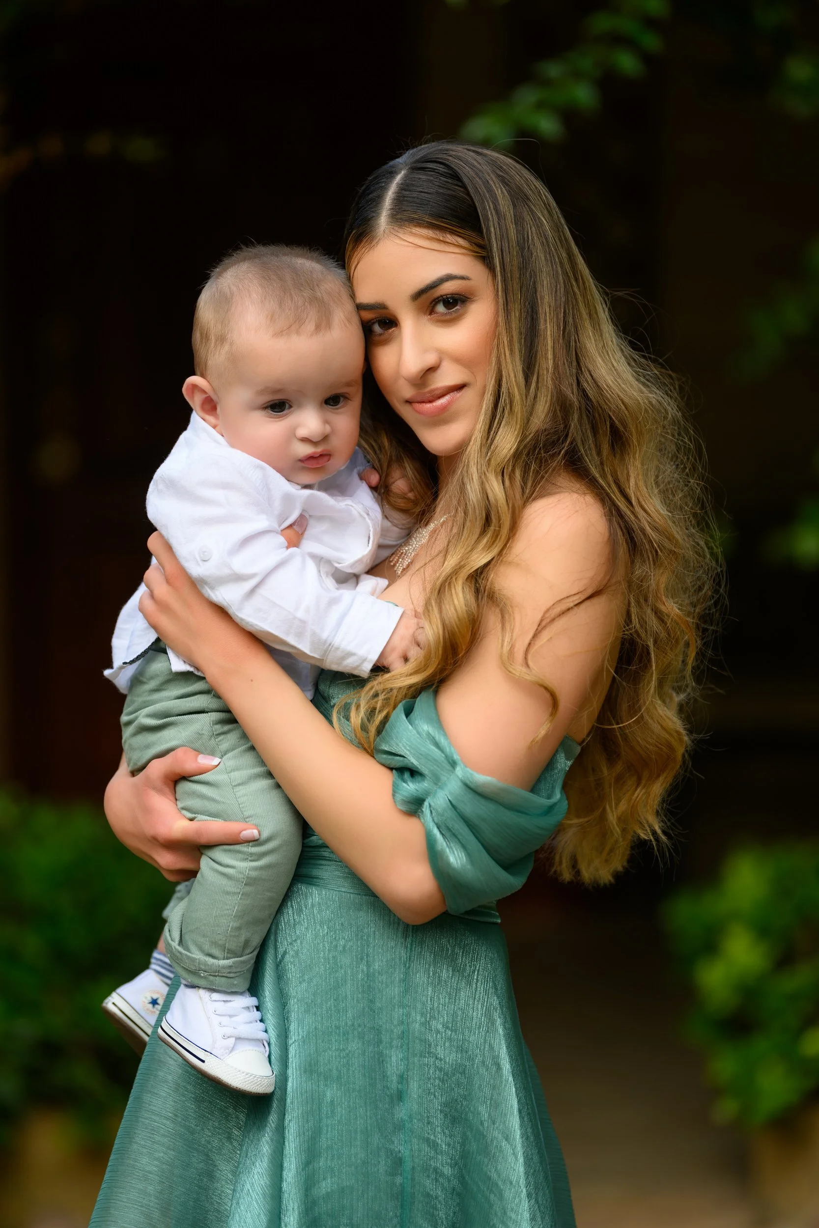 A woman with long, wavy, light brown hair holding a young child with short, light brown hair, outdoors with greenery background.