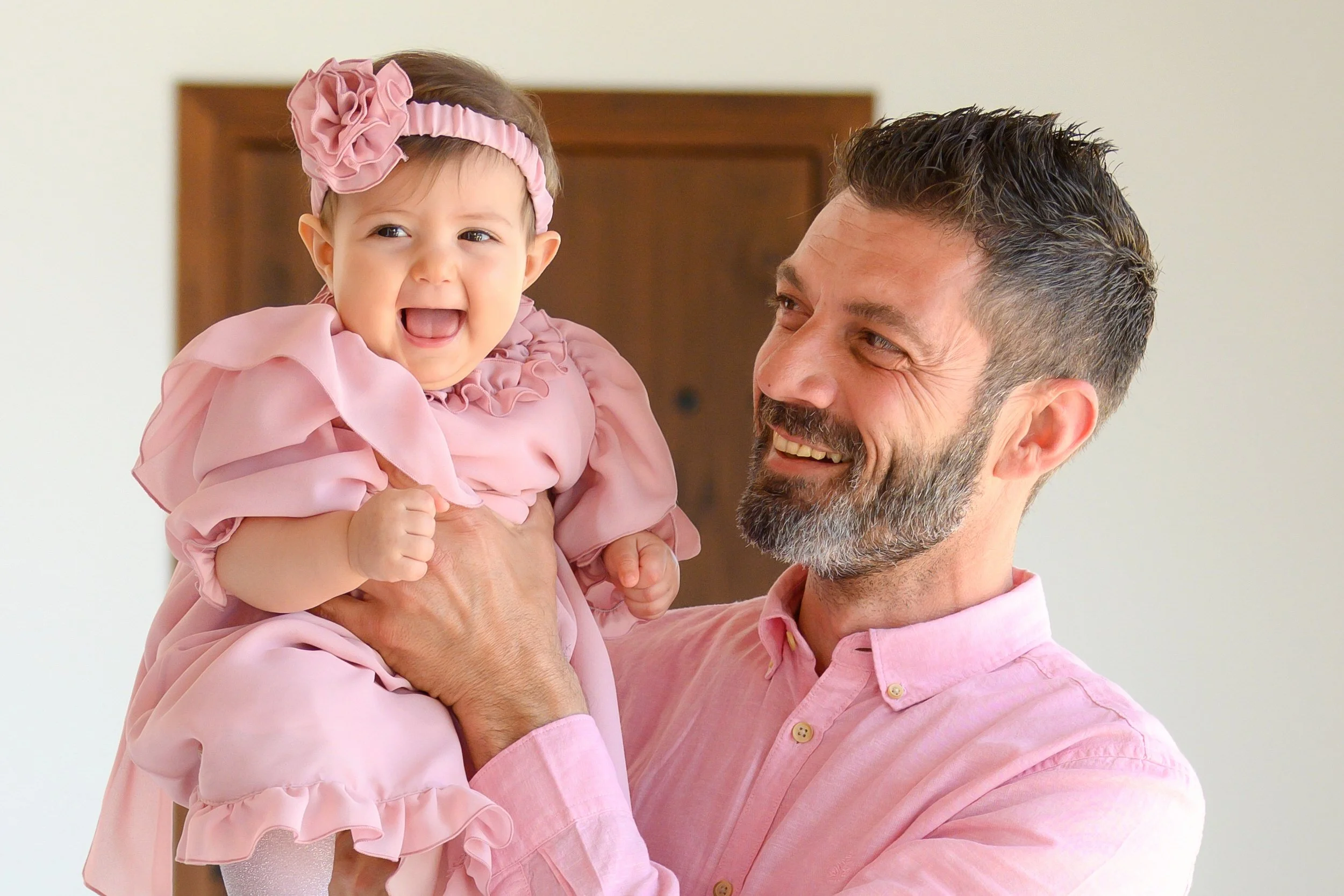 A man smiling and holding a laughing baby girl in a pink dress and headband.