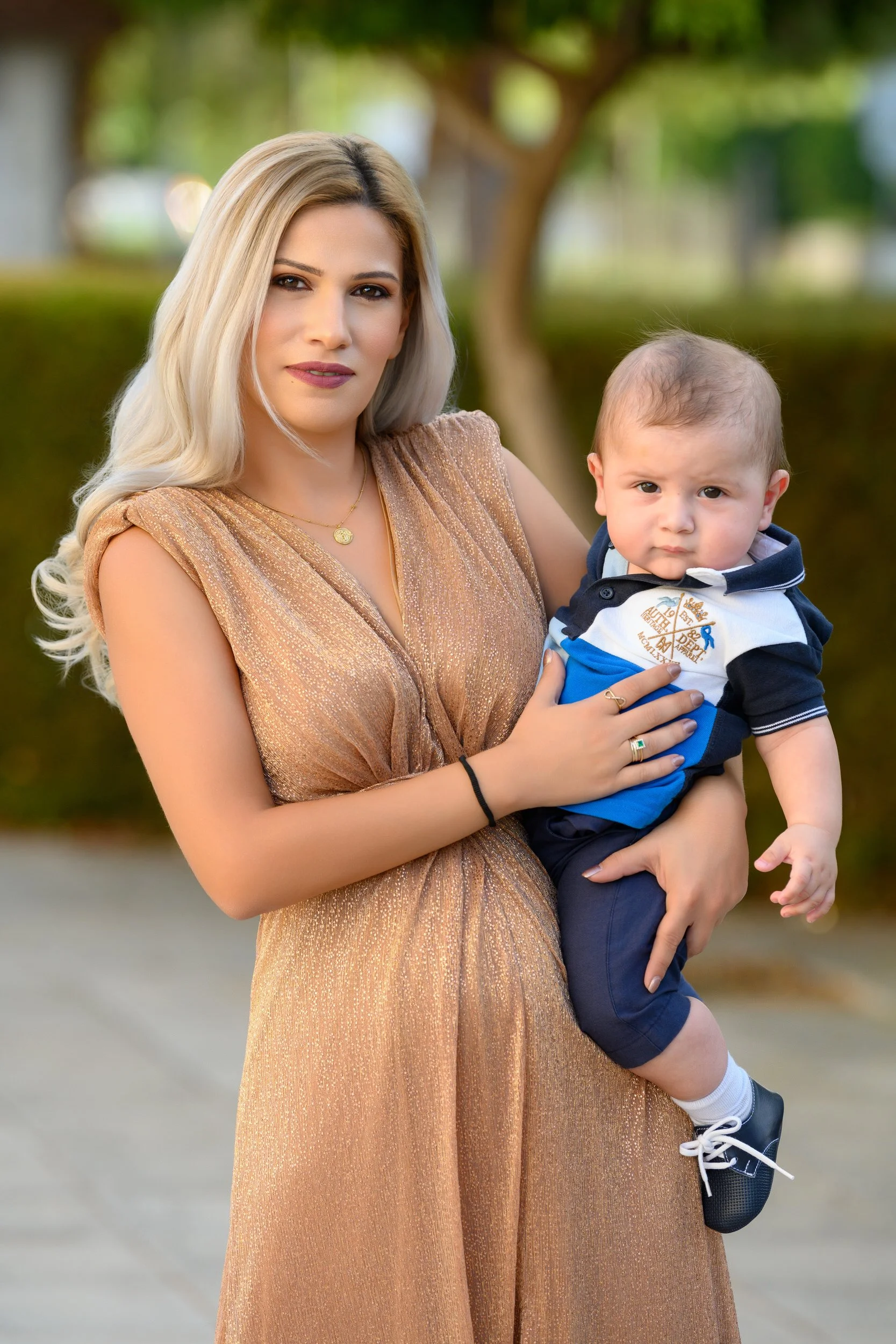 A woman with long blonde hair, dressed in a gold-colored dress, holds a young boy with short light brown hair, wearing a white and blue polo shirt and navy shorts, outdoors near trees and bushes.