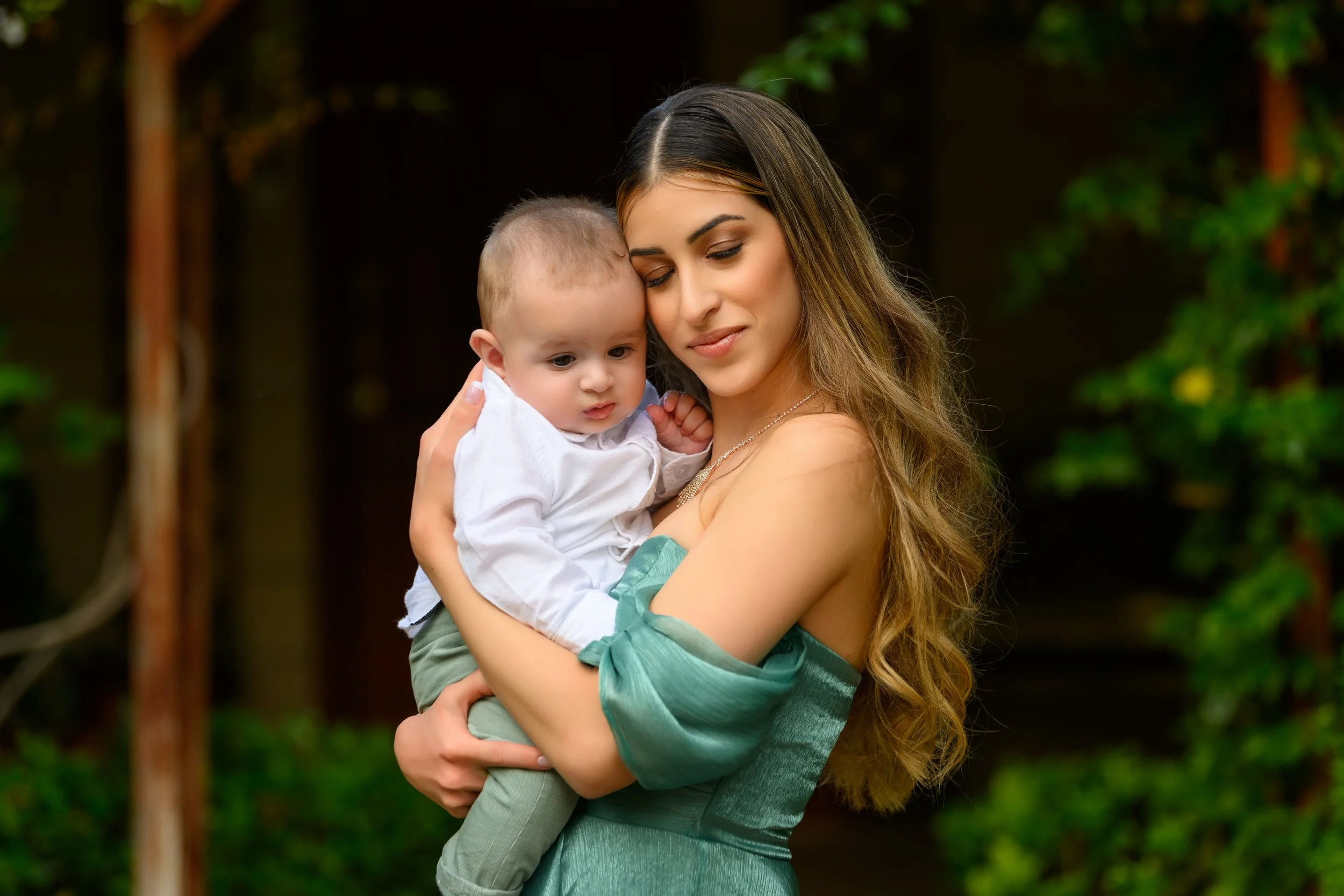 A woman with long wavy hair holding a young boy outdoors with trees in the background.