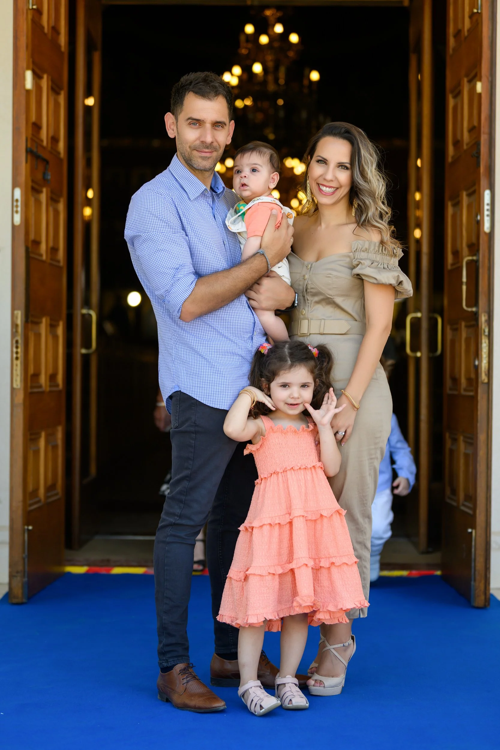 A family of four standing at an entrance, with a man in a blue checkered shirt holding a baby girl, a woman in a beige dress, and a young girl in a peach dress making a gesture with her hand.