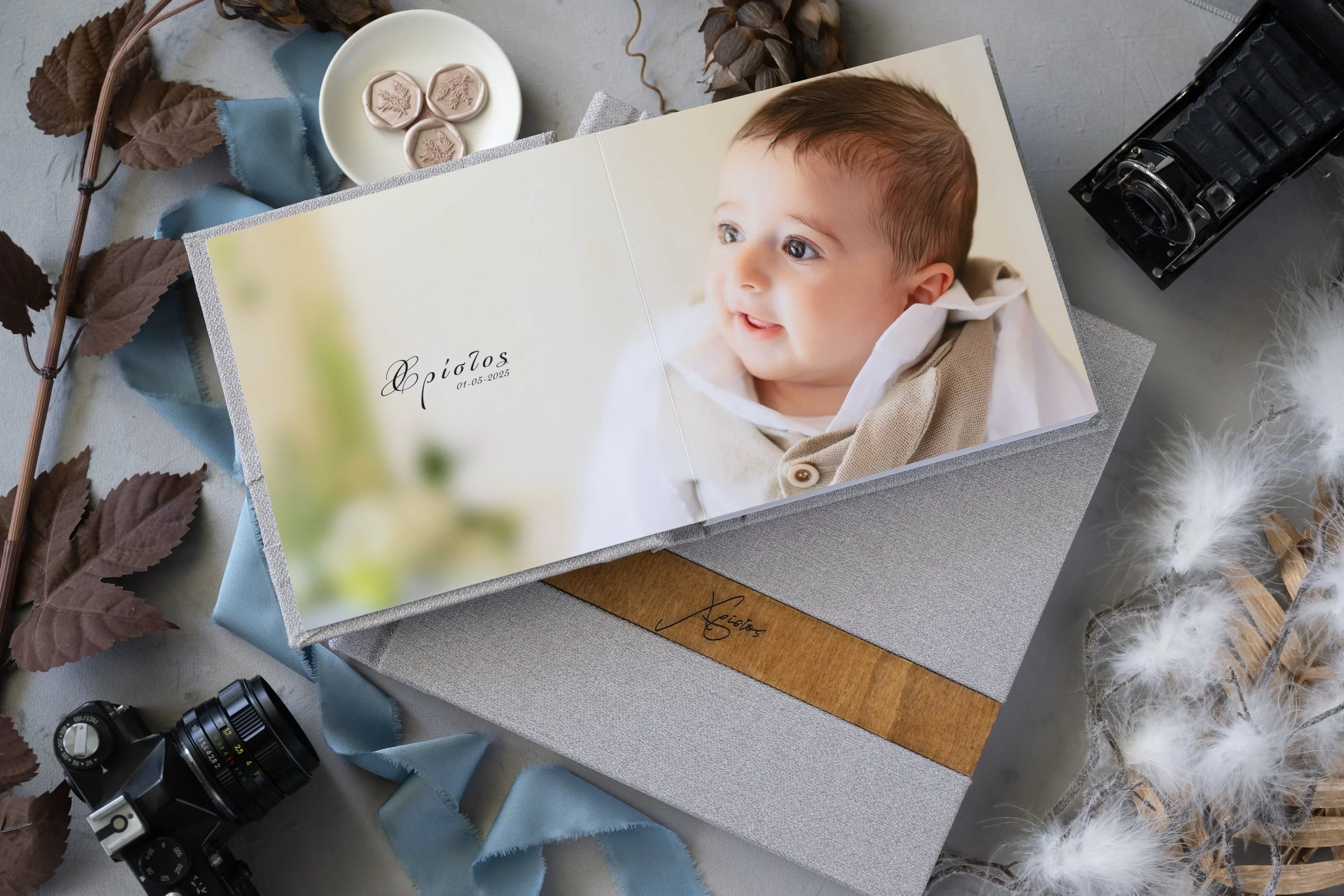 Open photo album with a picture of a smiling baby on a light-colored background. The album is placed on a textured surface surrounded by a vintage camera, a small bowl with wax seals, dried leaves, blue and white ribbons, and fluffy white feathers.