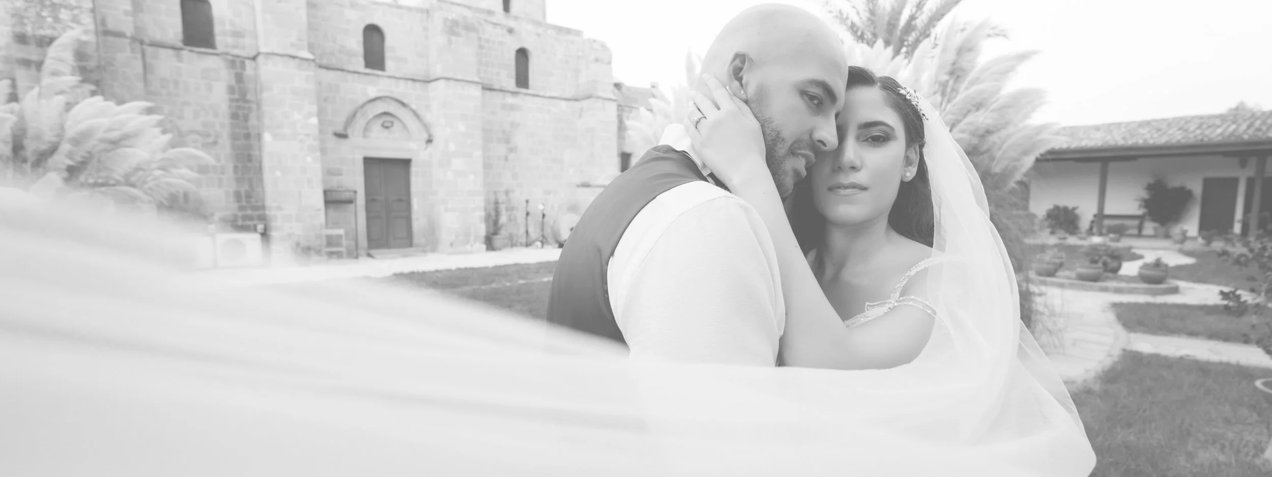 A black-and-white photo of a bride and groom embracing outdoors during a wedding, with a historic stone building and landscaping in the background.