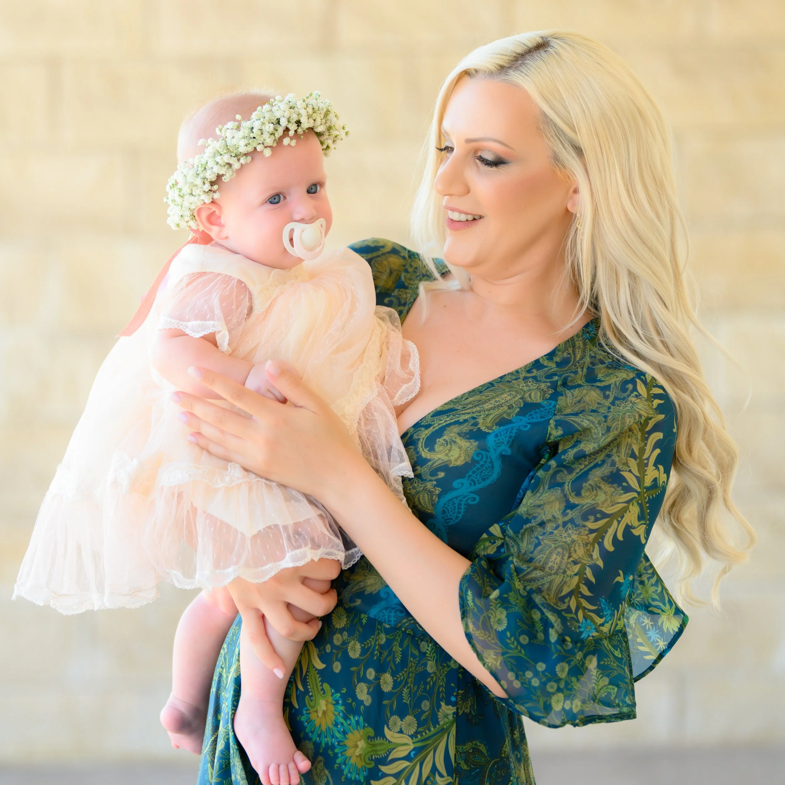 A smiling woman with long blonde hair holding a baby girl dressed in a peach-colored dress with a flower crown and pacifier, standing against a light background.