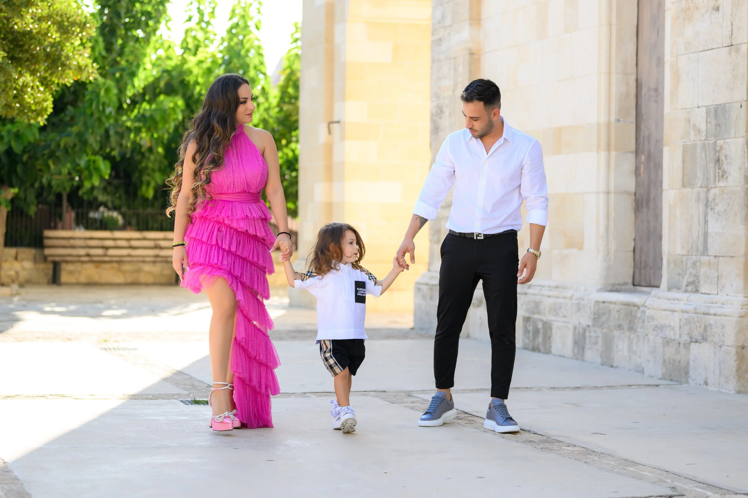 A family of four walking outdoors near a stone building and green trees, with the mother in a pink ruffled dress holding hands with a young girl, and the father holding the other hand of the girl.