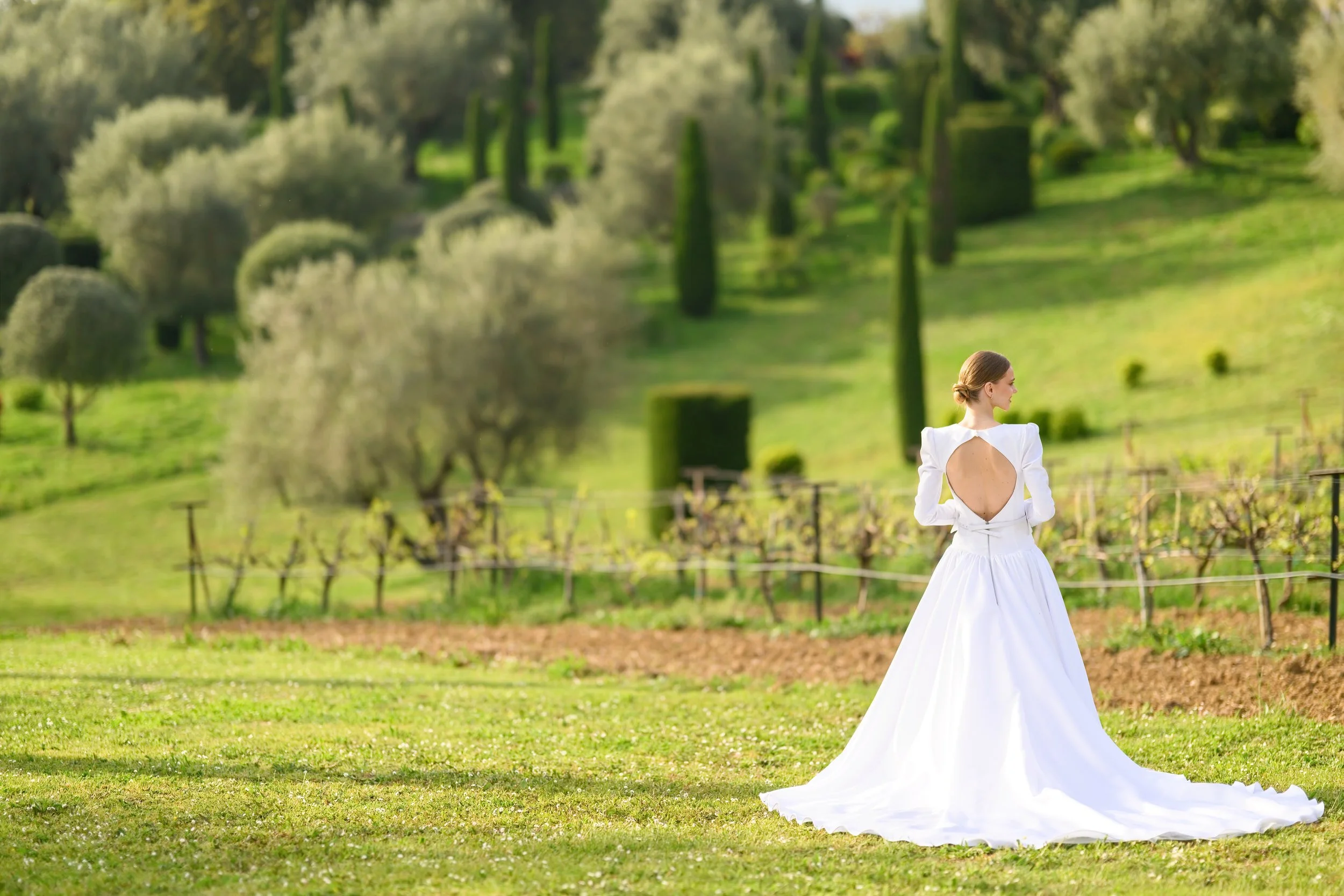 A woman in a white wedding dress standing in a lush green vineyard on a sunny day.