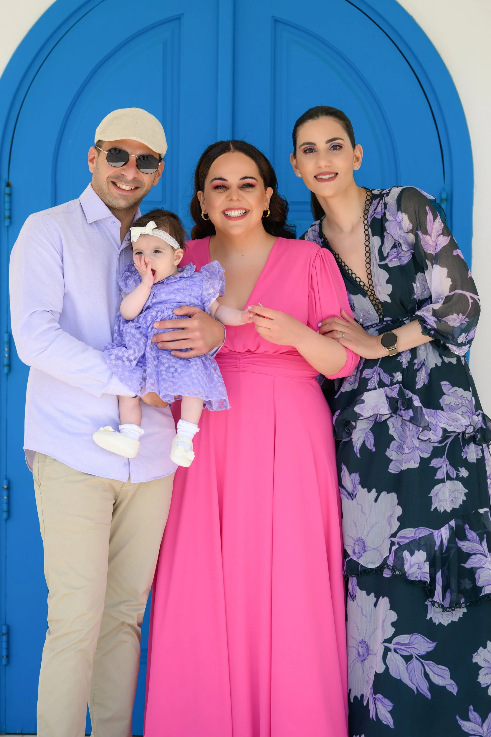 A family of four standing in front of a bright blue door, including a man in sunglasses and a baby girl in a purple dress, a woman in a pink dress, and another woman in a purple floral dress.