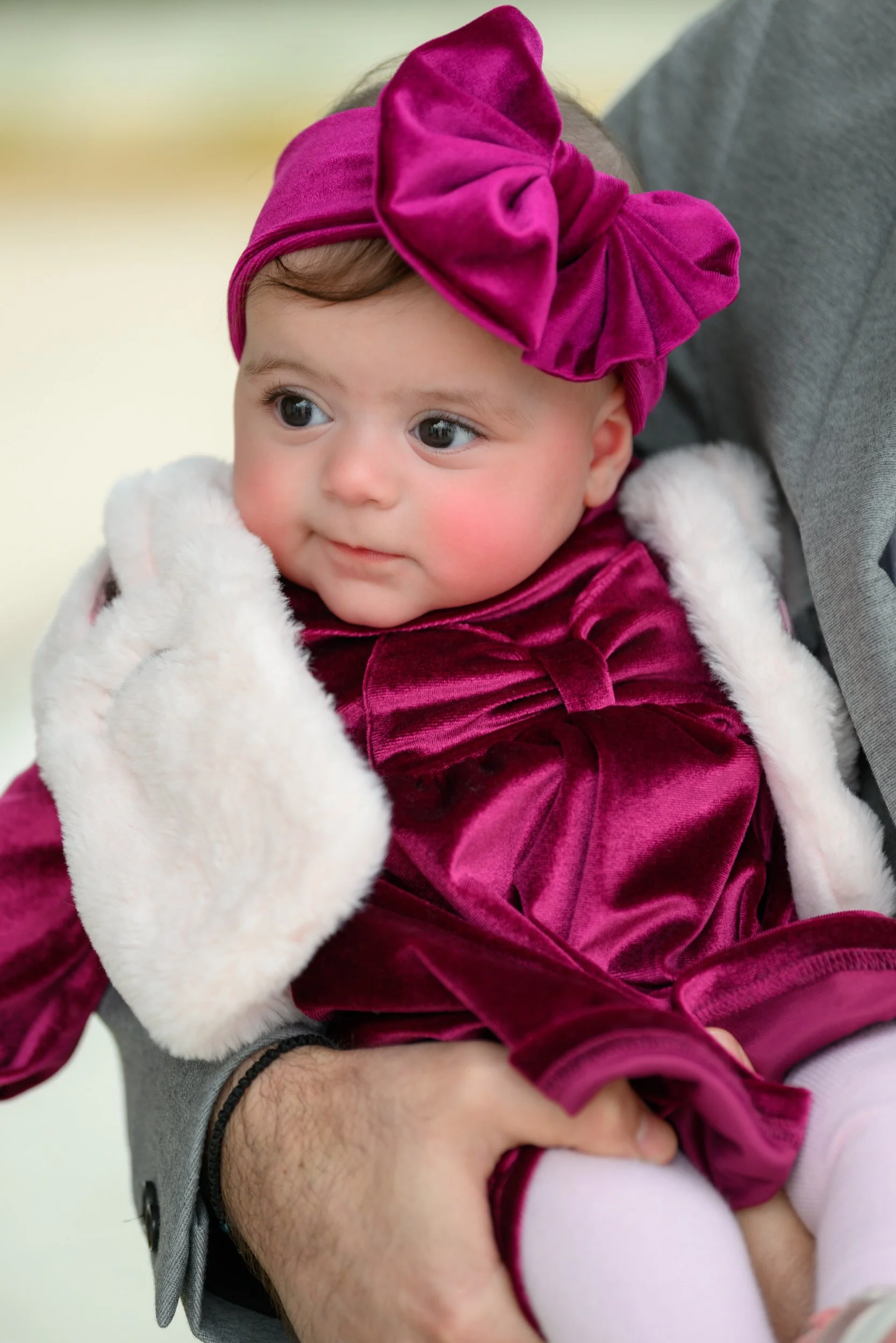 A baby girl wearing a maroon velvet dress and a matching headband with a large bow, sitting on an adult's lap, holding her face with one hand, and gazing to her right.