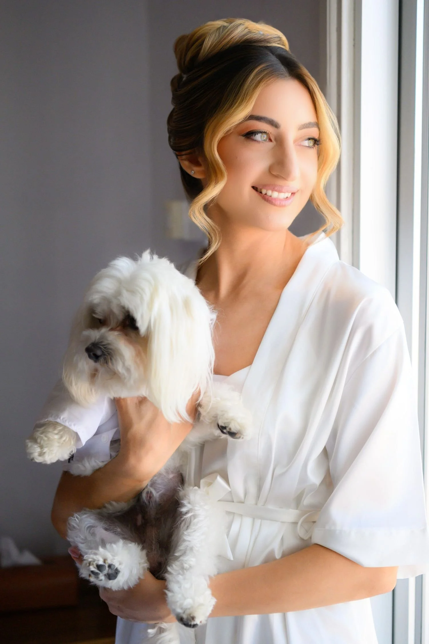 A woman in a white robe holding a small white and gray dog by a window.