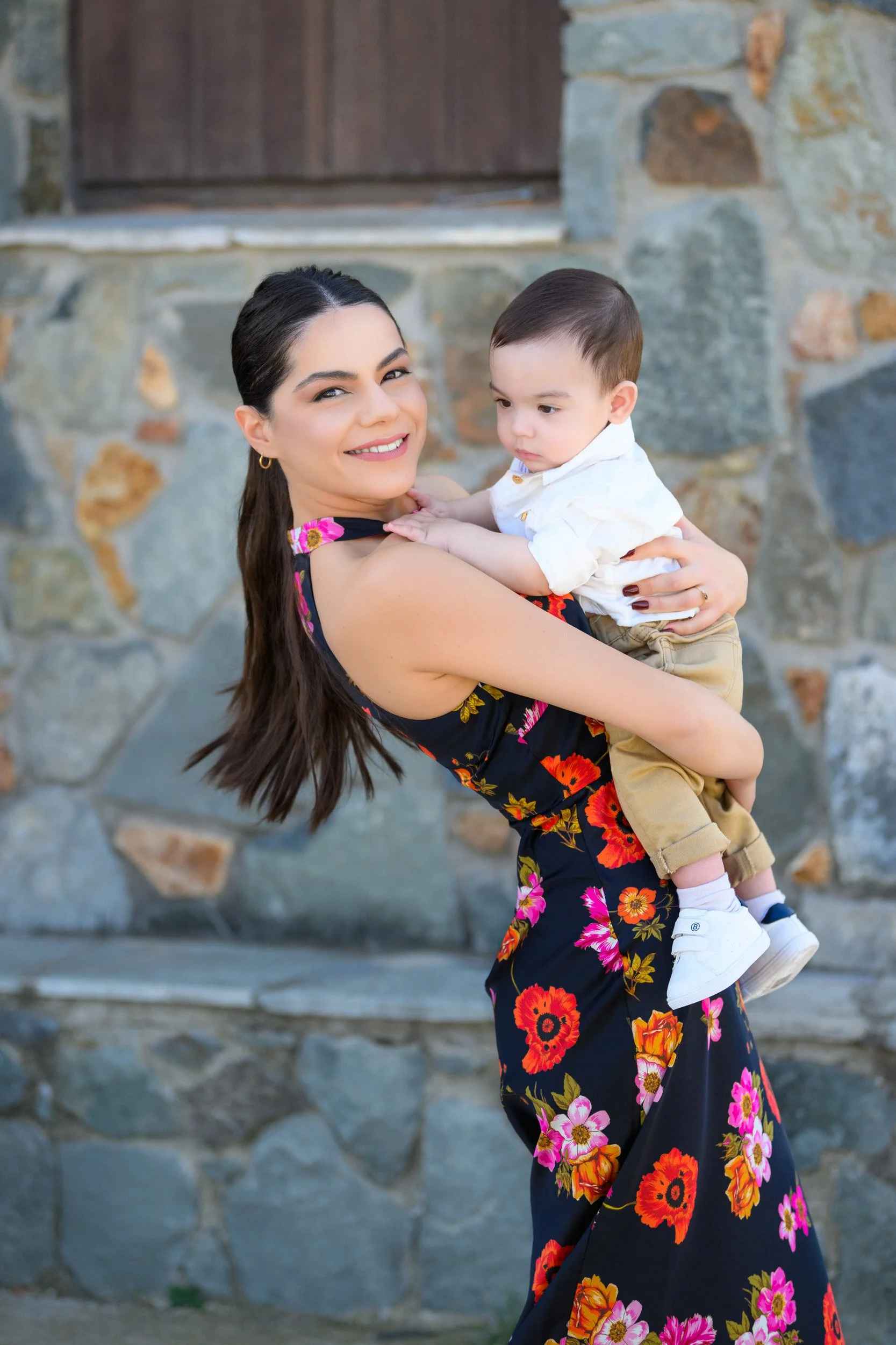 A woman with long dark hair, wearing a black floral dress, smiling while holding a young boy in front of a stone wall and wooden door.