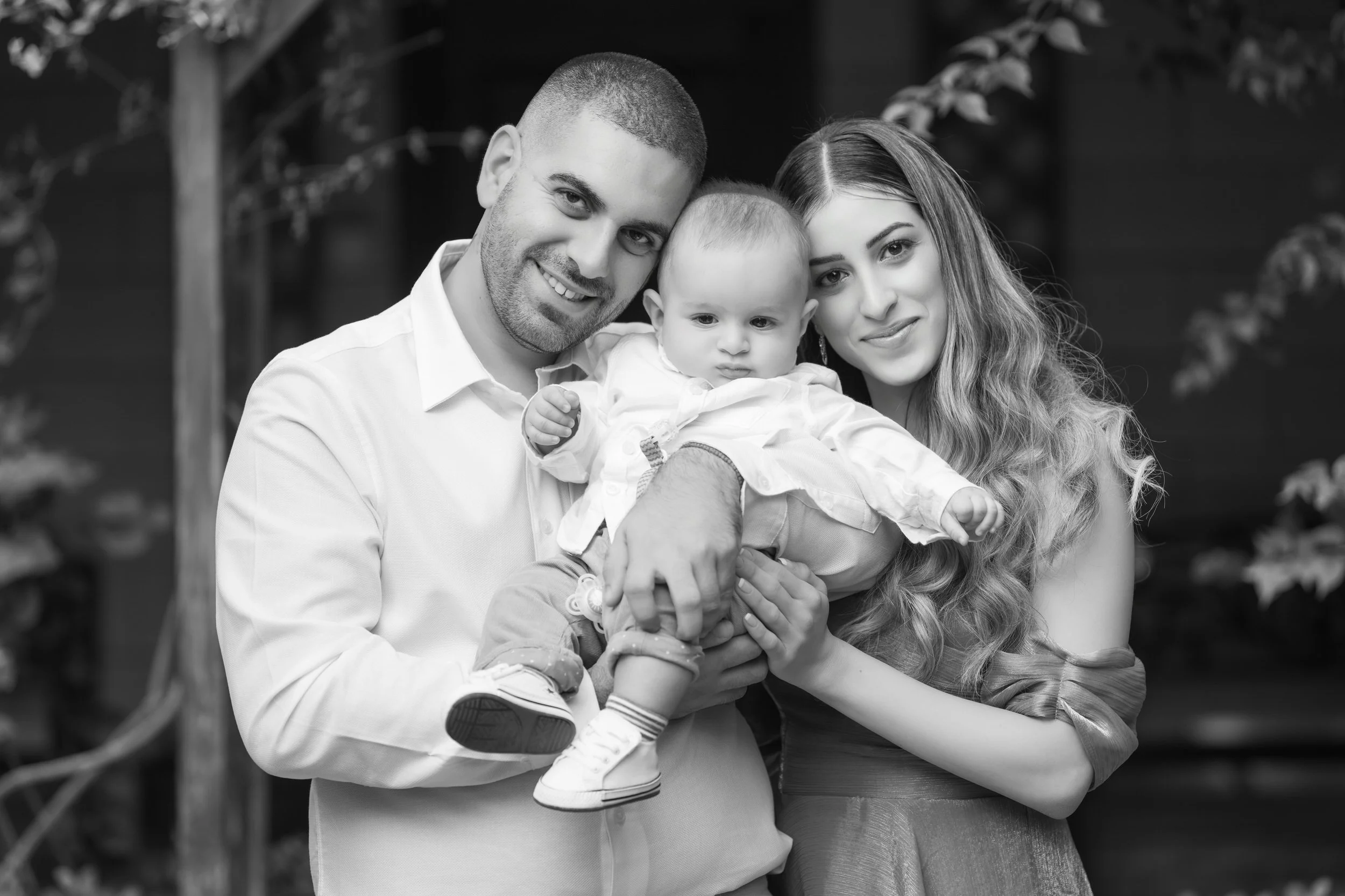 A happy family of three, with a man, a woman, and a baby, posing together outdoors. The man is holding the baby, and all three are smiling at the camera.