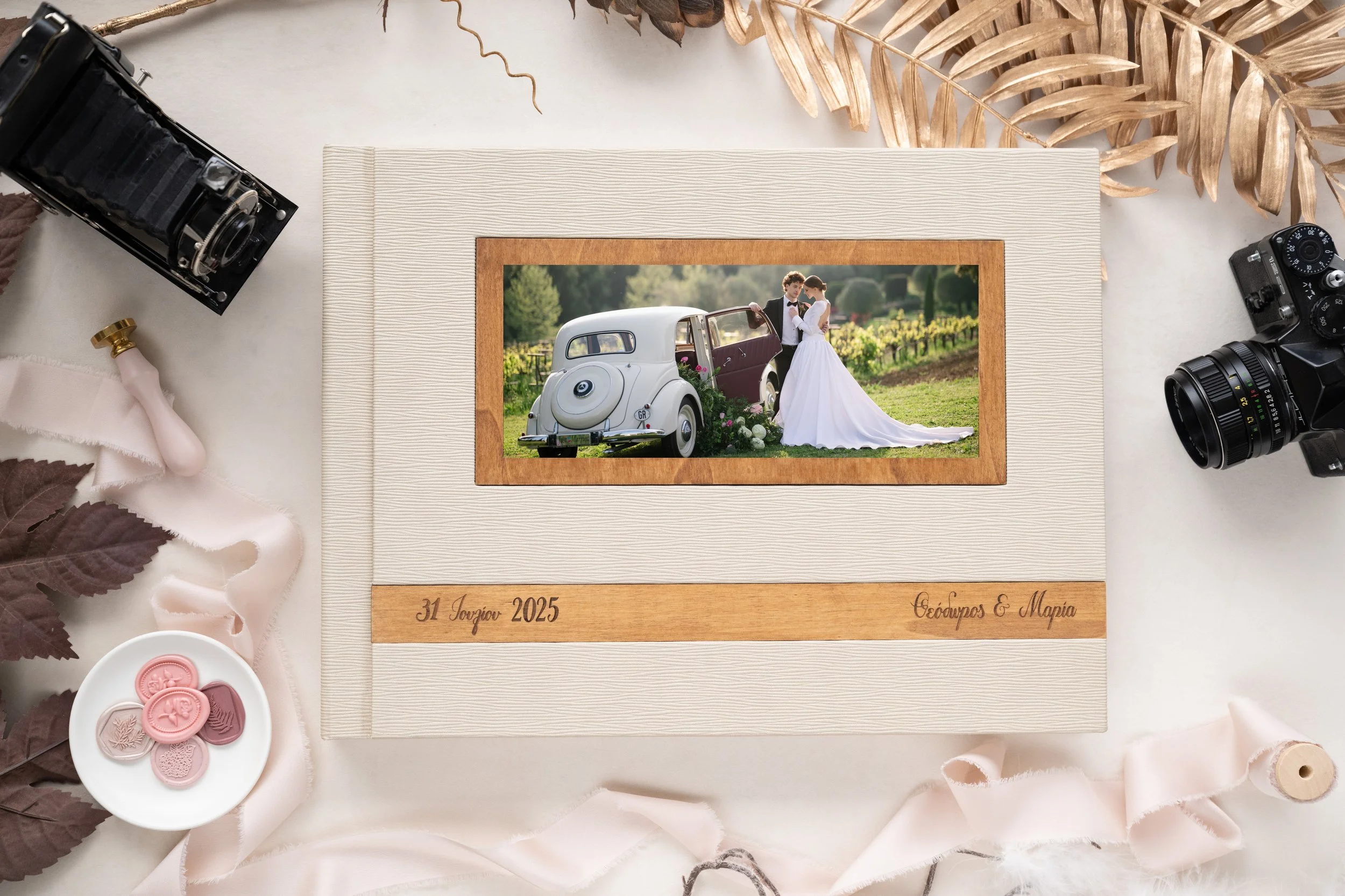 A wedding photo album on a table surrounded by vintage cameras, ribbons, leaves, and pink wax seals, with a picture of a bride and groom standing next to a vintage car in a vineyard setting, engraved with the wedding date '31 July 2025' and the names