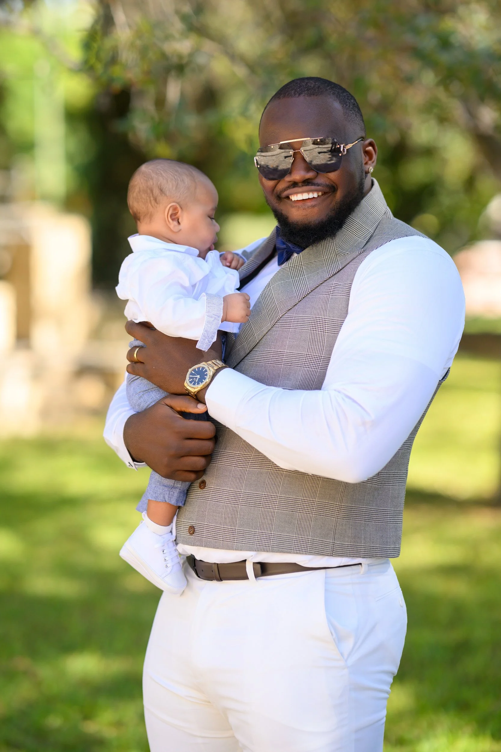 A man in sunglasses, a gray vest, white shirt, and white pants holding a young child outdoors in a park, smiling at the camera with green trees in the background.