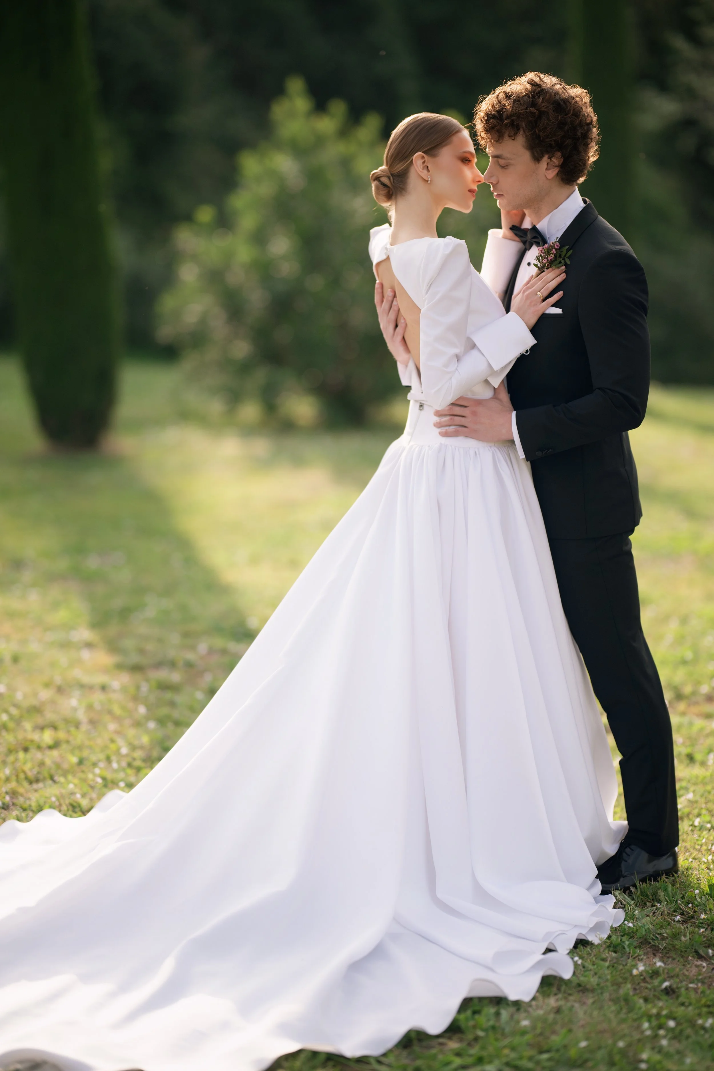 Bride and groom holding each other outdoors in a park setting, dressed in wedding attire, with the bride in a white gown and the groom in a black suit, facing each other.