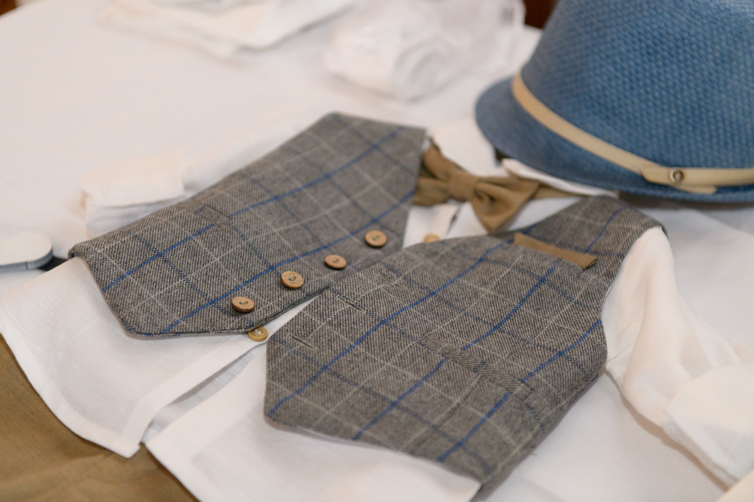A gray plaid vest with buttons, a white shirt underneath, and a brown bow tie. There is a blue hat nearby on a white surface.