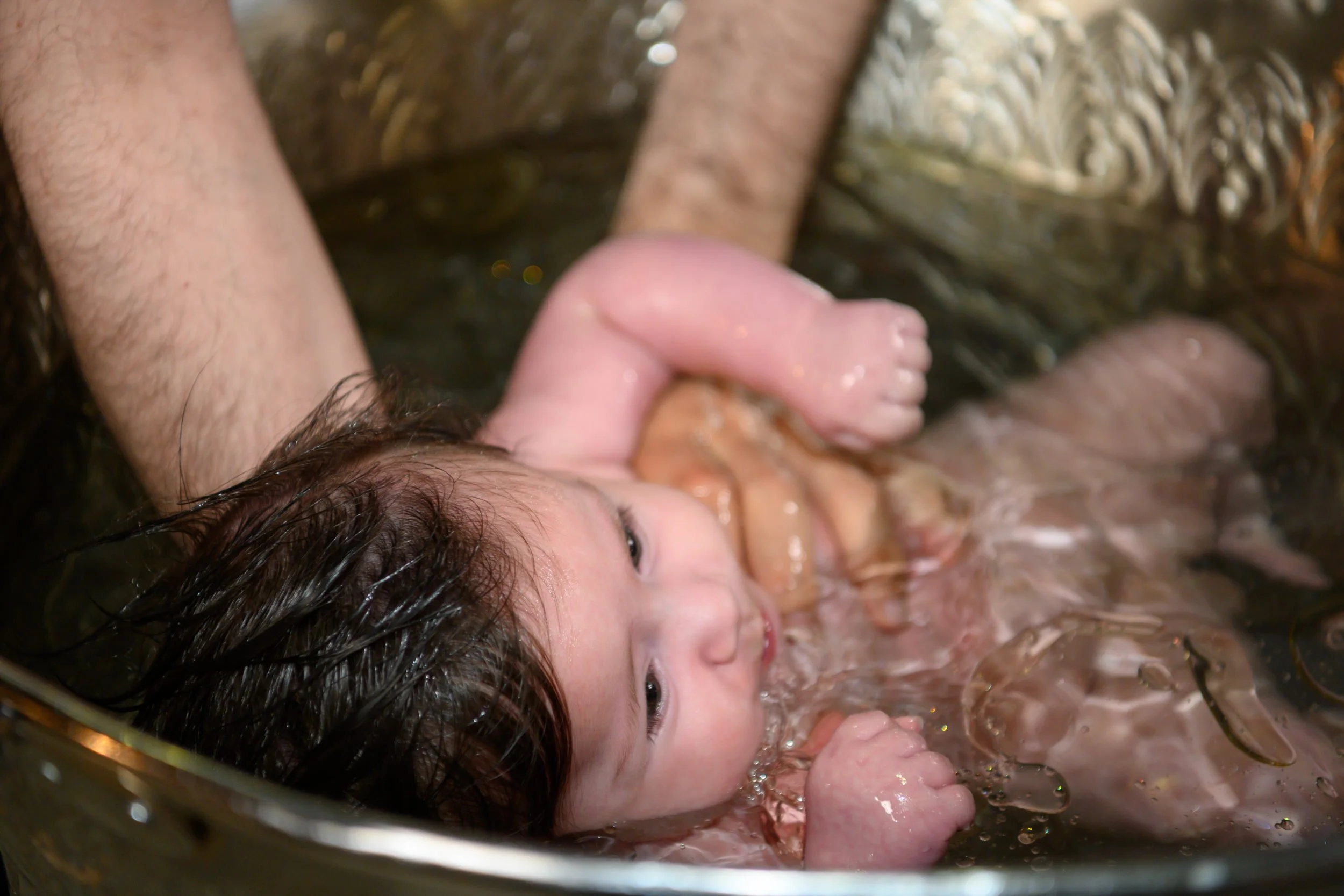 A young child with wet hair being baptized in a metal tub by an adult.