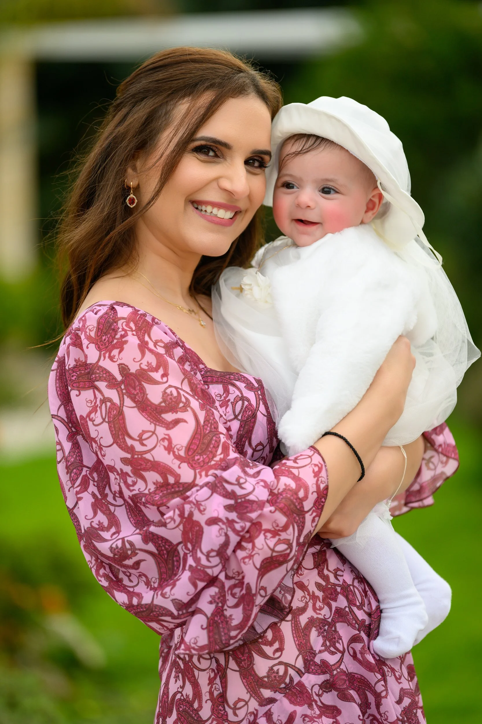 A woman holding a smiling baby dressed in white outdoors with green blurred background.