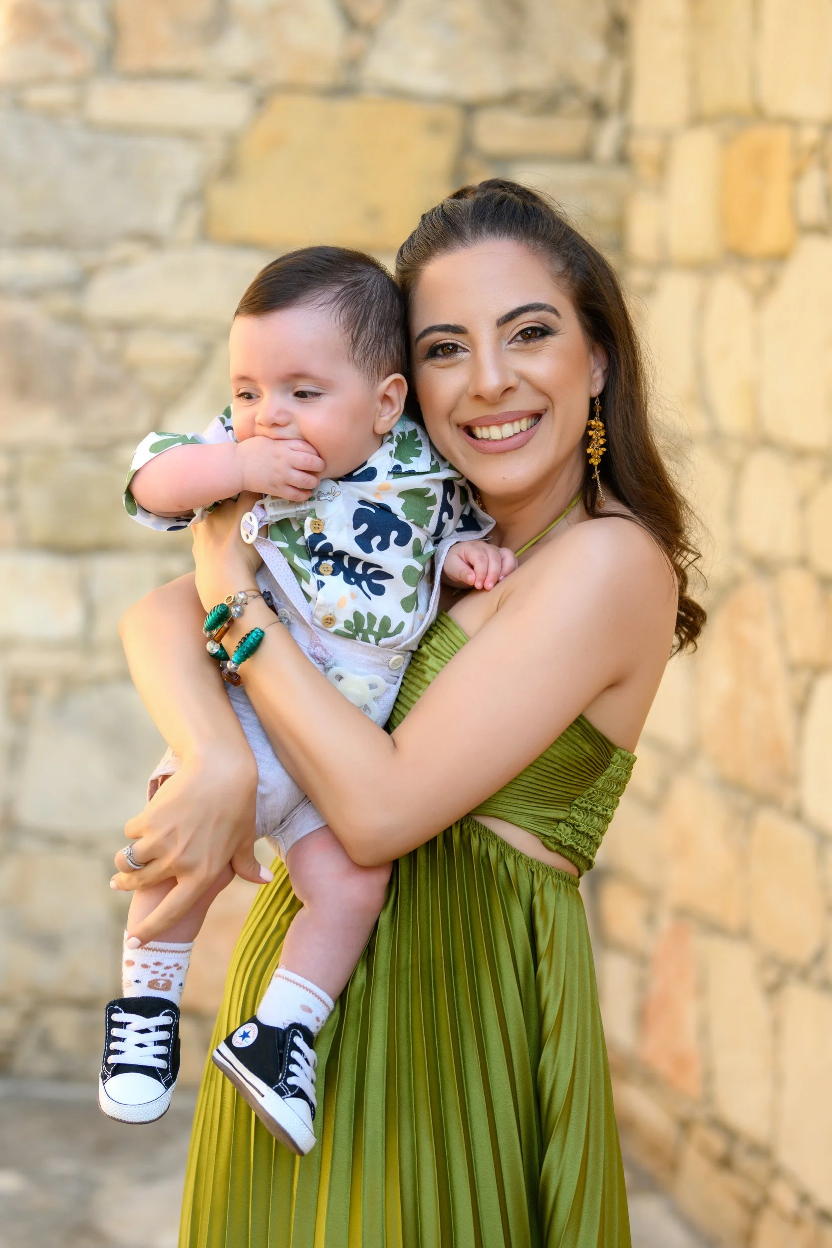 A woman holding a young boy outdoors, smiling, with a stone wall in the background.