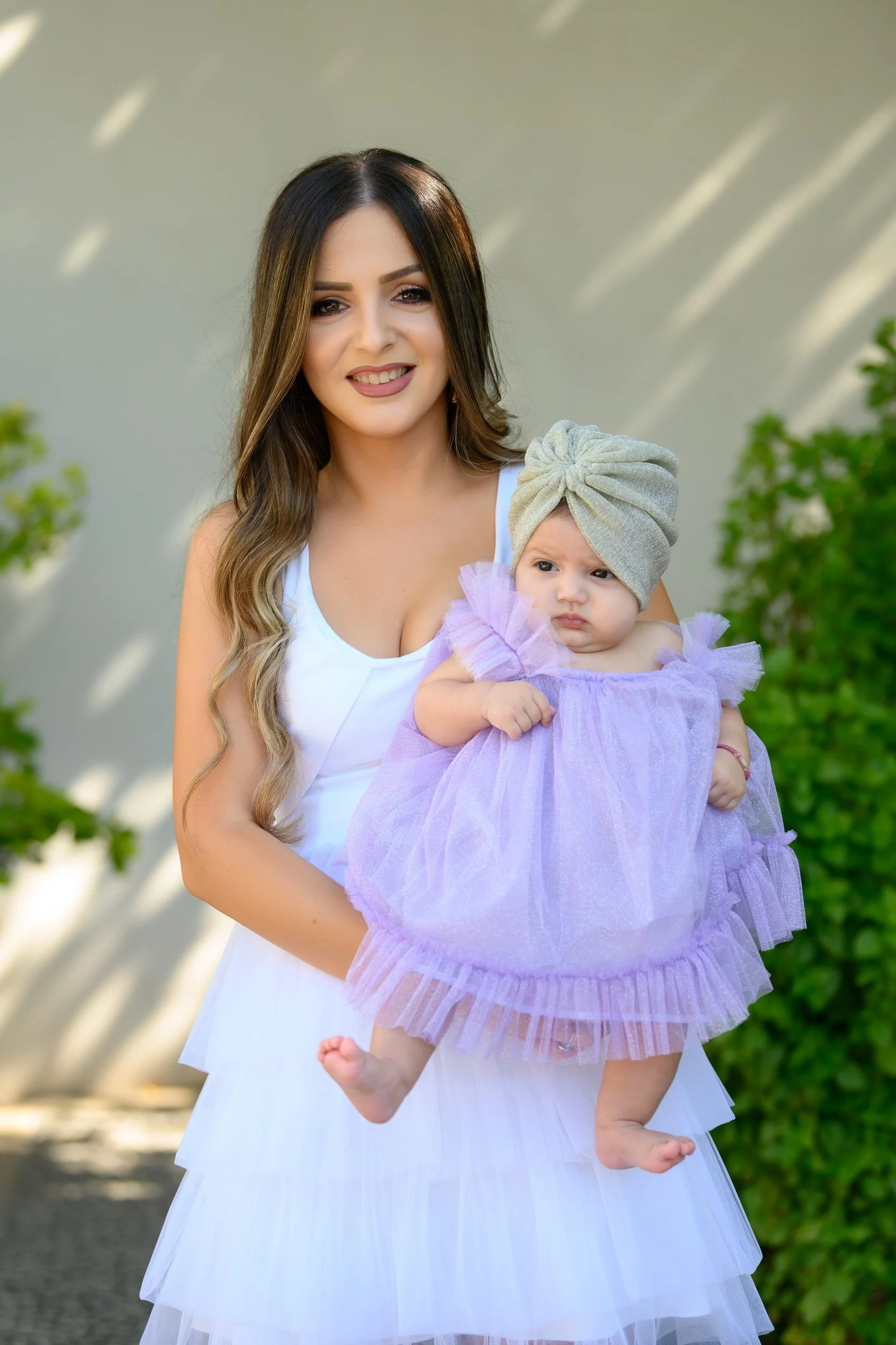 A woman holding a young girl outdoors during daytime, with both facing the camera. The woman has long wavy hair and is wearing a white dress. The young girl is dressed in a purple tutu dress and has a gray turban-style headband.