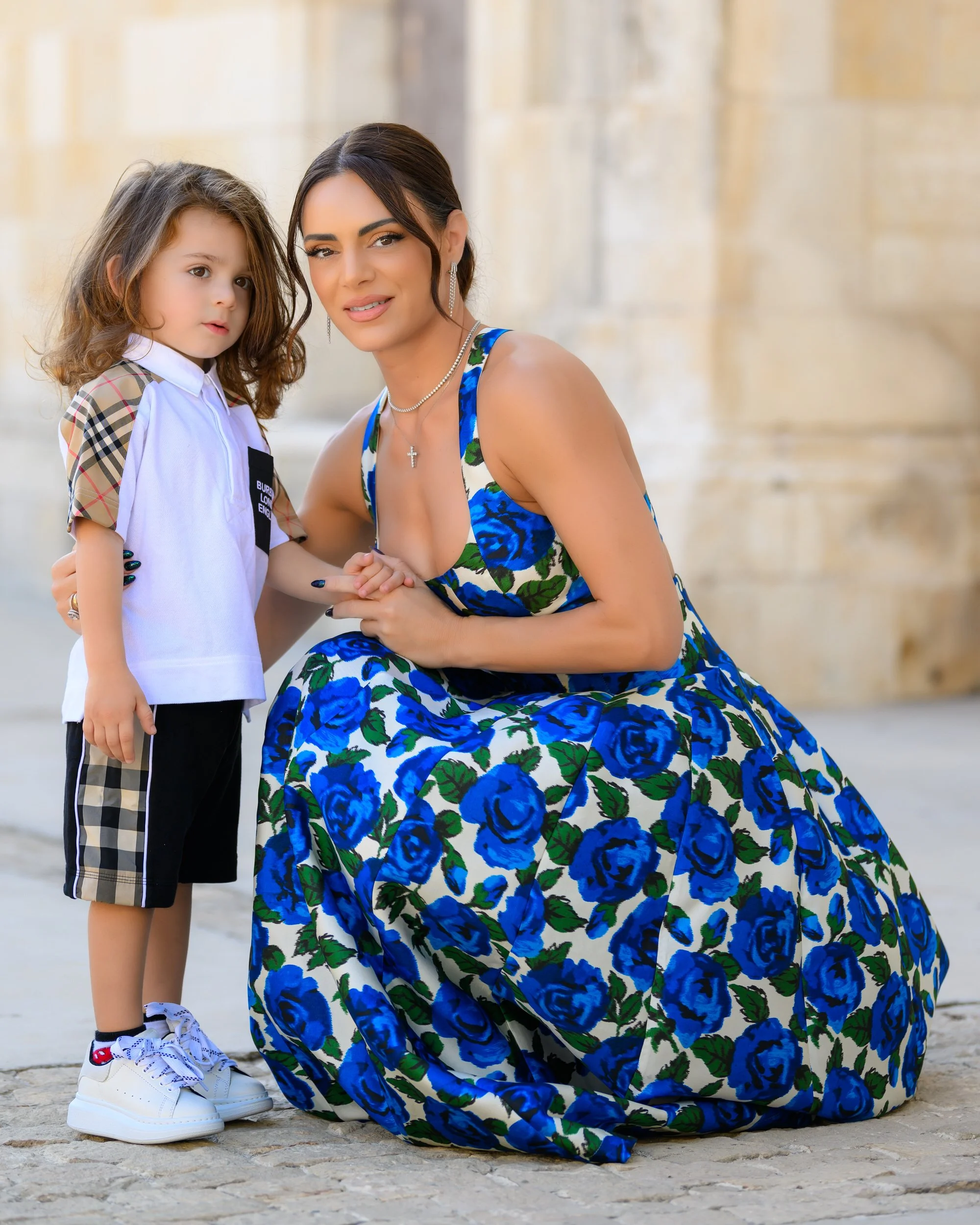 A woman in a blue floral dress kneeling beside a young girl in a soccer uniform, holding hands, outside in front of a stone wall.