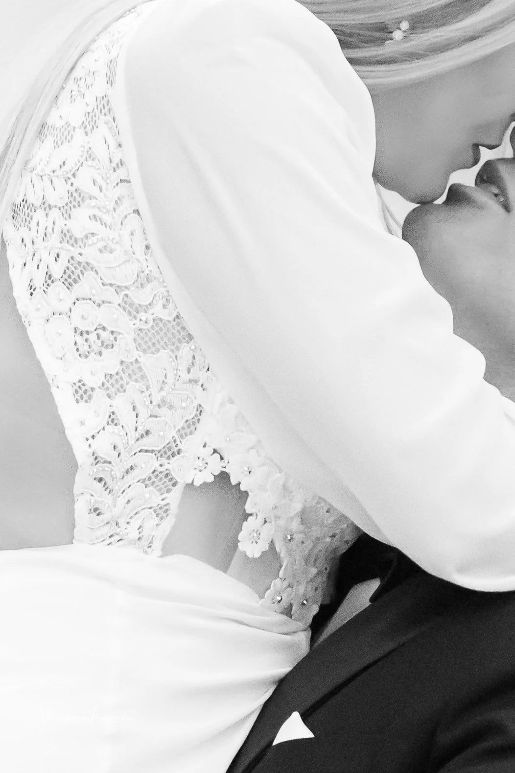 A close-up black-and-white photo of a bride and groom kissing. The bride's dress features detailed lace on the sleeves and bodice, and the groom is wearing a dark suit with a white shirt and pocket square.