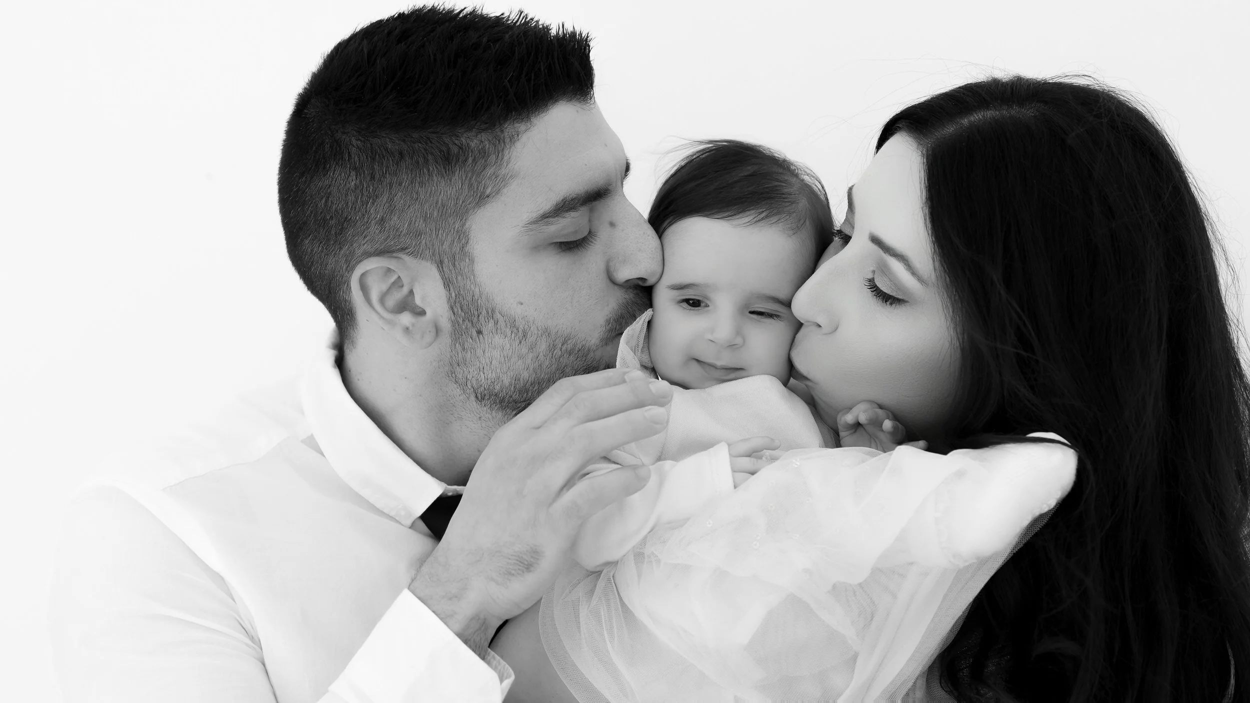 Black and white photo of a happy couple holding their baby, with the man and woman kissing the baby's cheeks, showing love and tenderness.