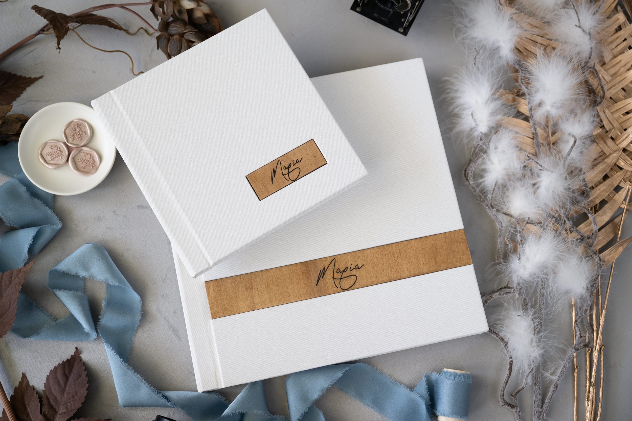 Close-up of two white photo albums with brown leather labels that have the name 'Maria' written on them, placed on a grey surface surrounded by dried leaves, blue ribbon, a small white dish with wax seals, and a bundle of white fluffy decorative feat