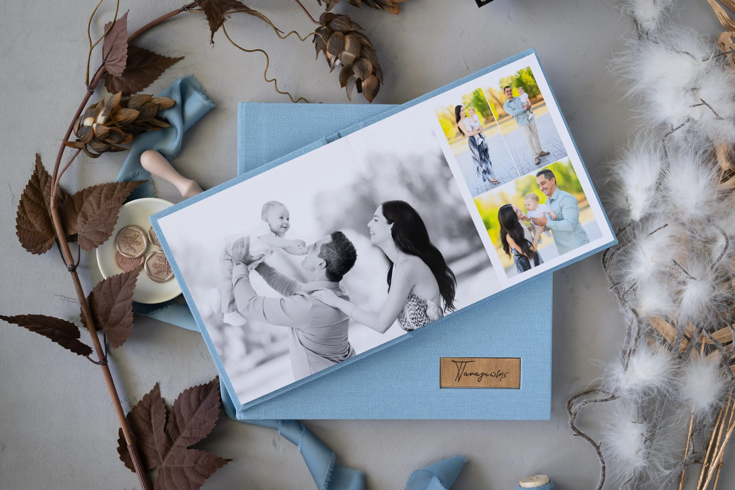 Open photo album with family pictures, surrounded by dried leaves, a ribbon, feathers, and wax seals.