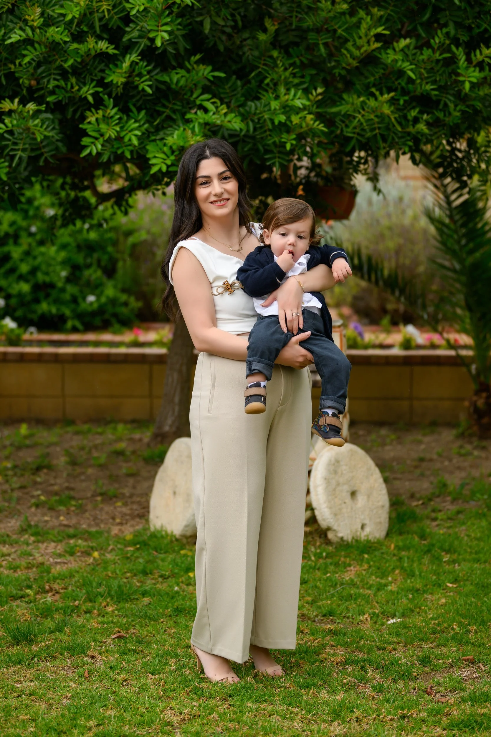A woman with long dark hair holding a young child outdoors in a garden with trees and greenery.