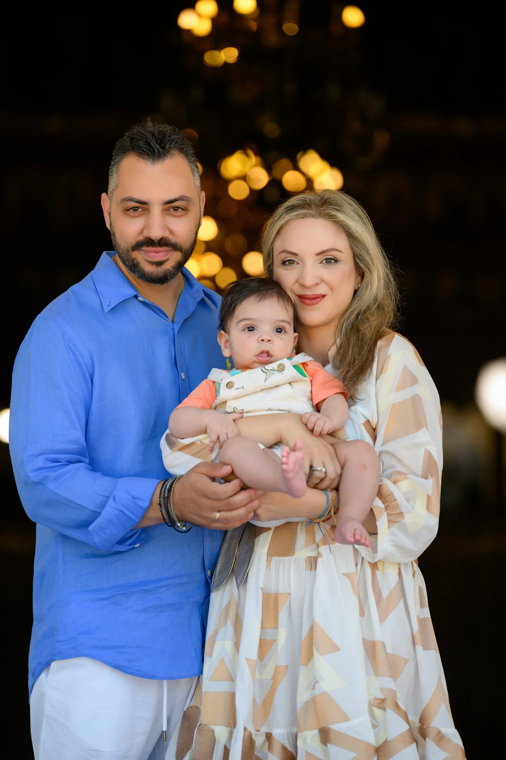 A family of three, a man, a woman, and a baby girl, posing together with blurred golden lights in the background.