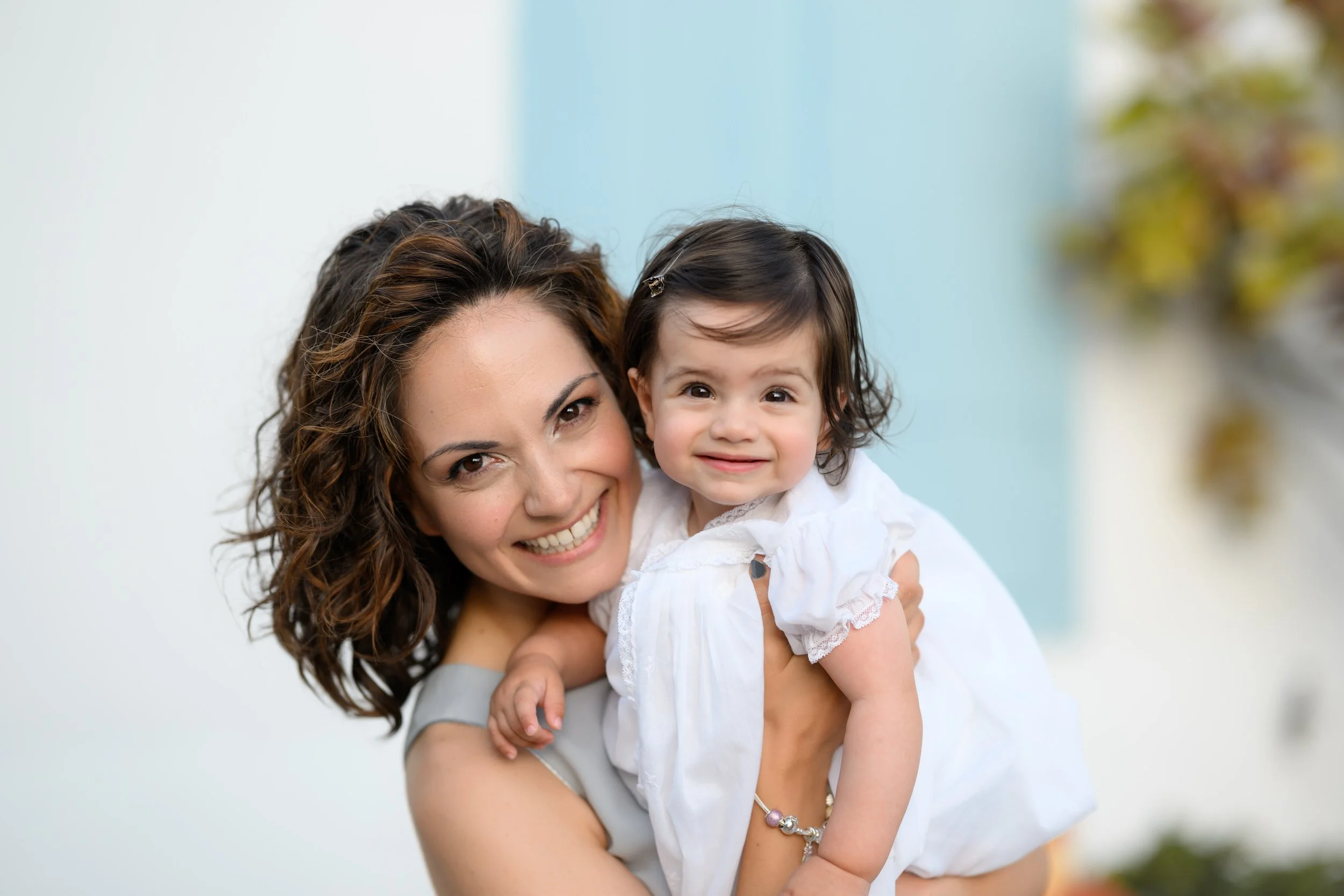 A woman with curly brown hair smiling and holding a young girl with dark hair and a white dress, both looking at the camera.