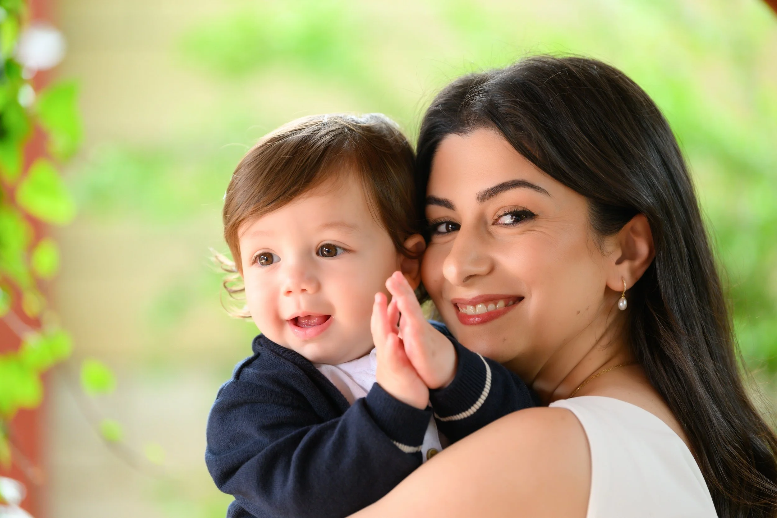 A woman holding a young child outdoors with a blurred green background, both smiling and looking at the camera.