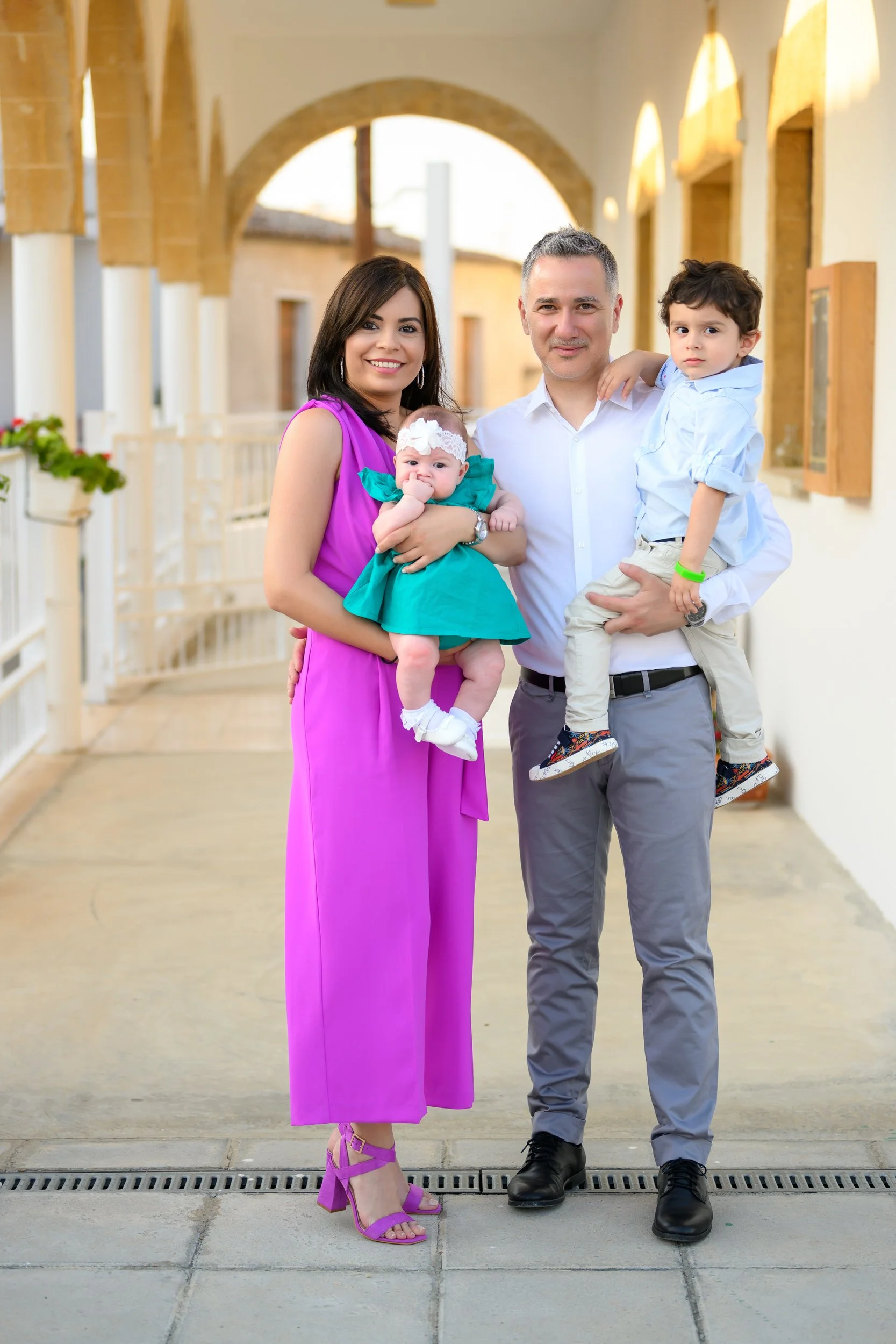 A family of four standing outside under an archway, smiling and posing for the photo. The woman is wearing a bright pink dress with matching pink heels and holding a baby girl in a green dress. The man is wearing a white shirt and gray pants, holding